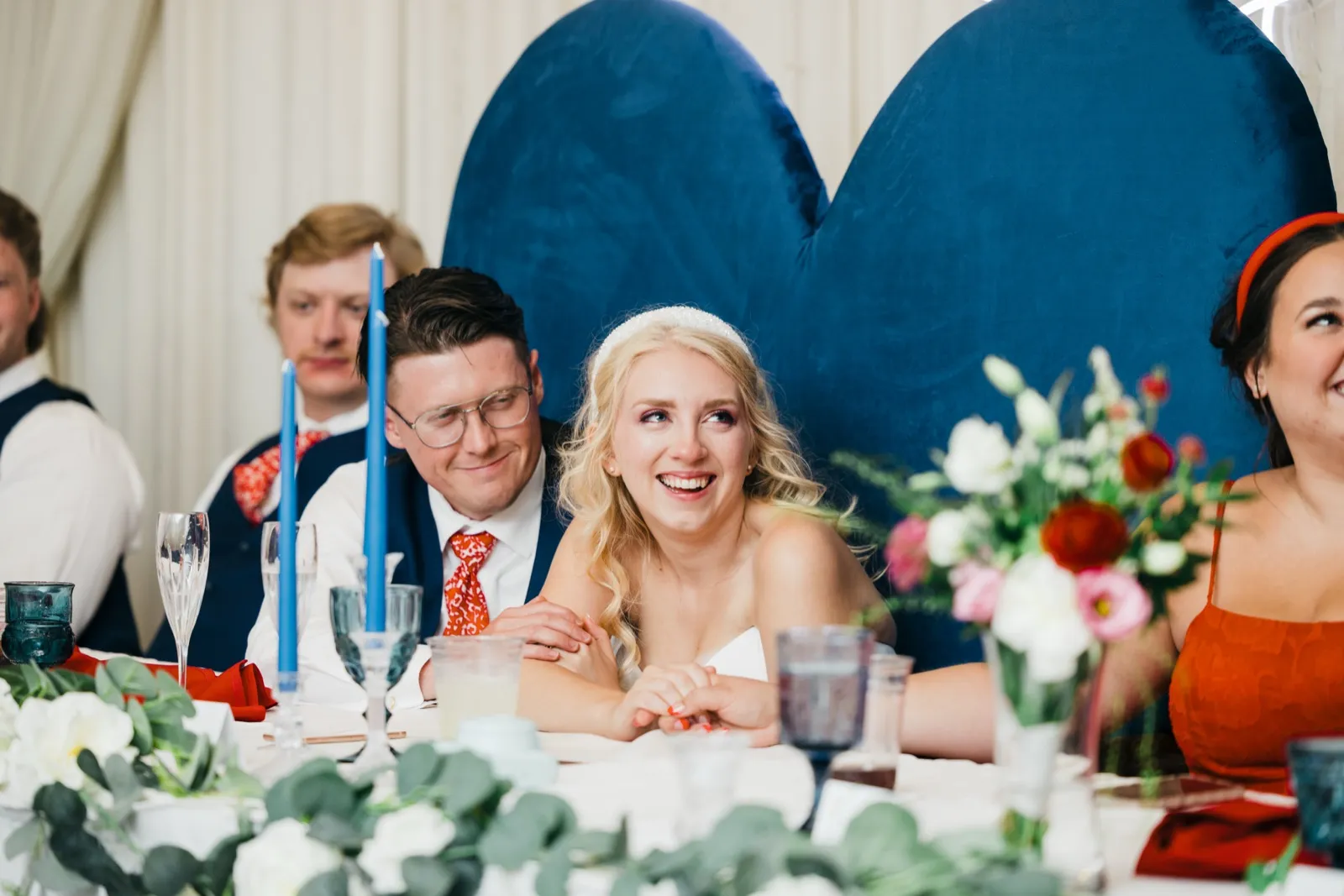 The bride laughs with her head tilted as the groom watches her affectionately at the head table, framed by tall blue candles and a velvet heart backdrop. — Tim Larsen Photography, Brainerd Lakes MN