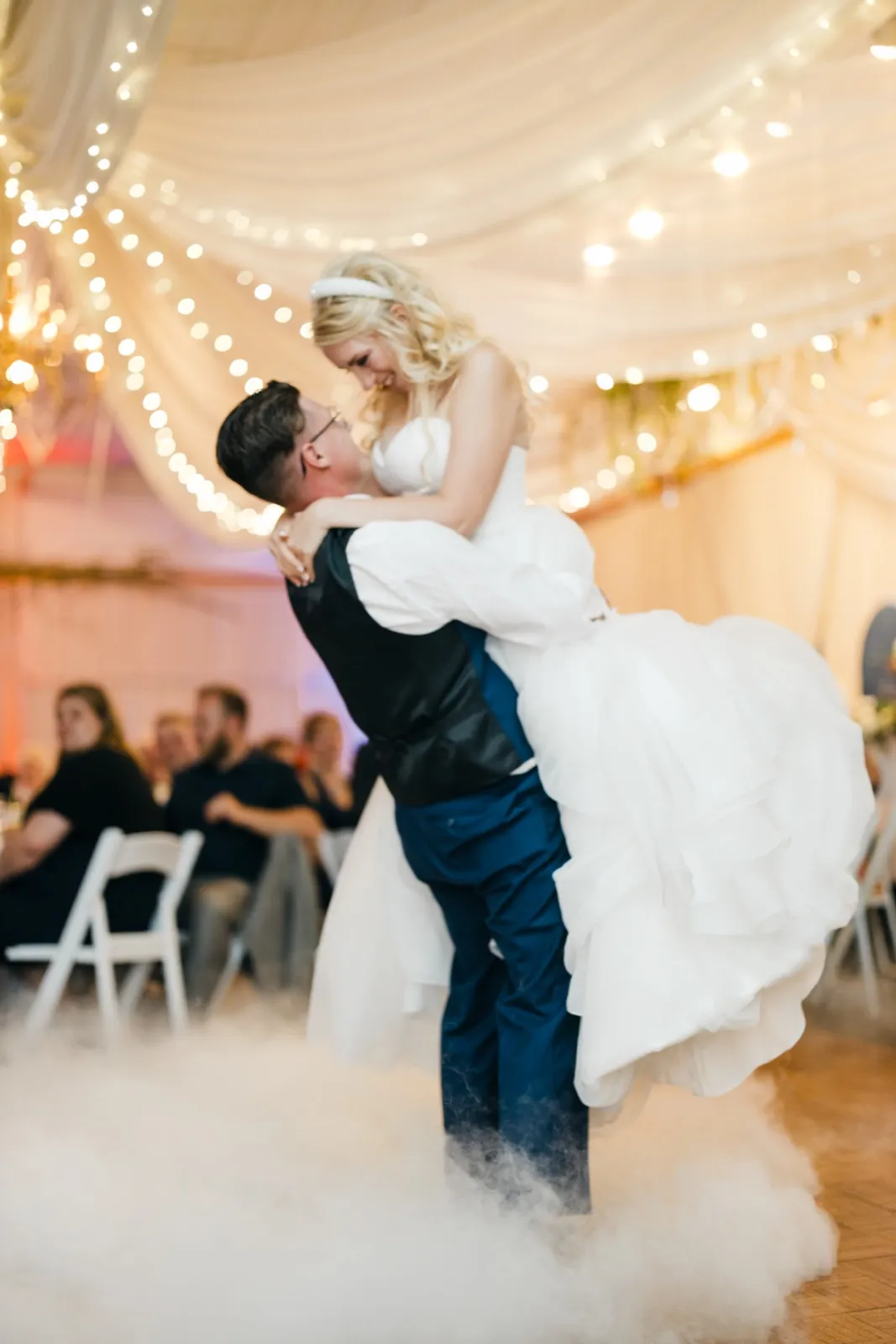 The groom lifts the bride off the floor during their first dance under a draped ceiling of string lights, with low fog rolling around their feet and seated guests watching in the background. — Tim Larsen Photography, Brainerd Lakes MN
