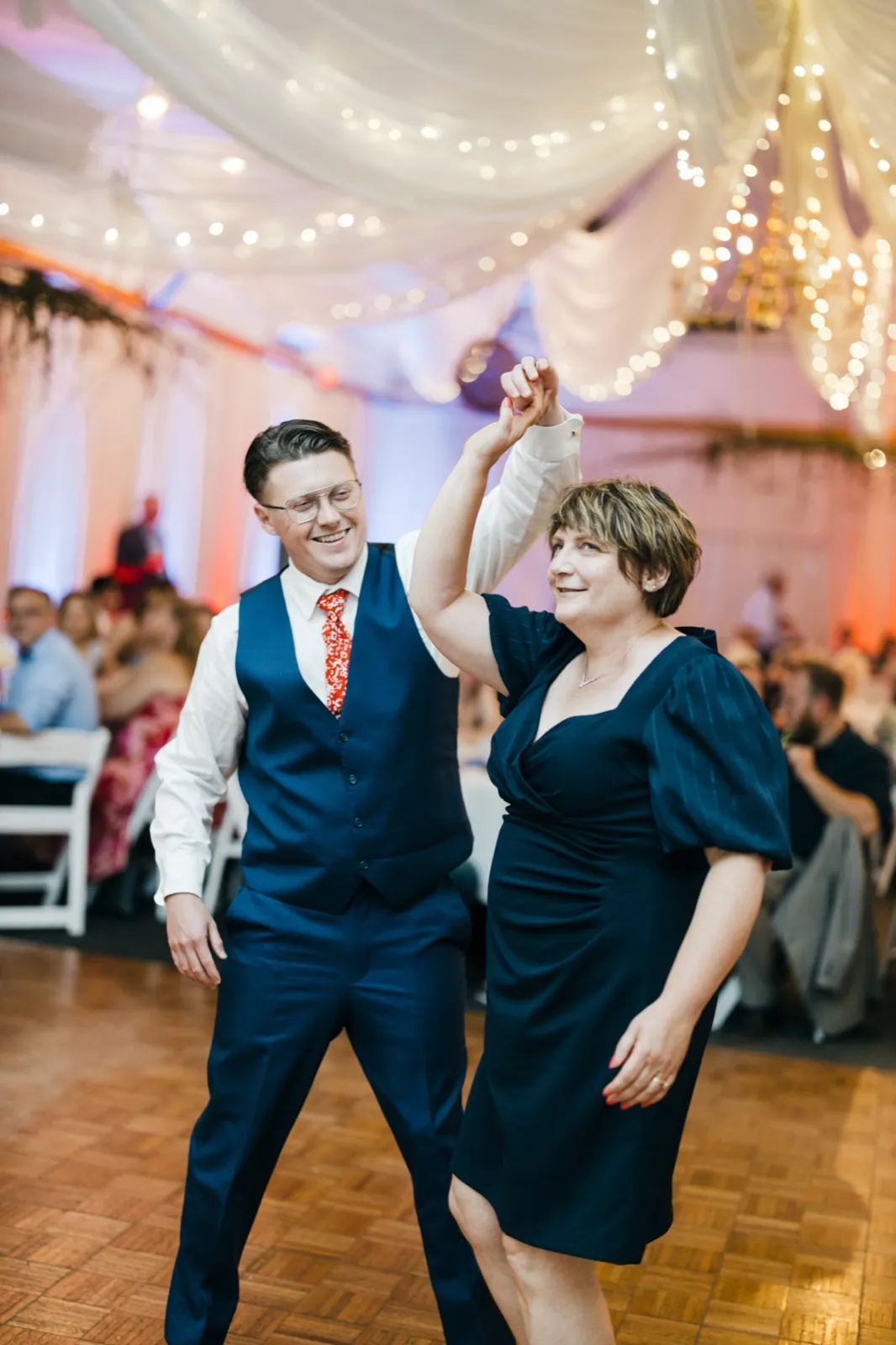 The groom twirls his mother on the dance floor beneath draped fabric and chandelier string lights at Pine Peaks Event Center. — Tim Larsen Photography, Brainerd Lakes MN