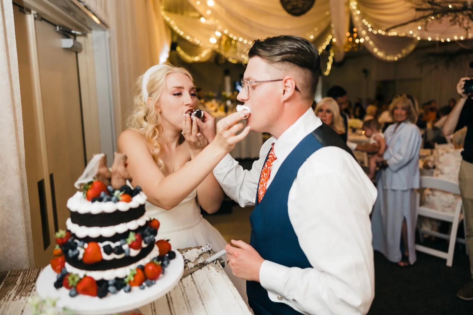 The bride and groom simultaneously feed each other bites of berry-topped naked cake at the reception, with guests visible behind them under string lights and draped fabric. — Tim Larsen Photography, Brainerd Lakes MN