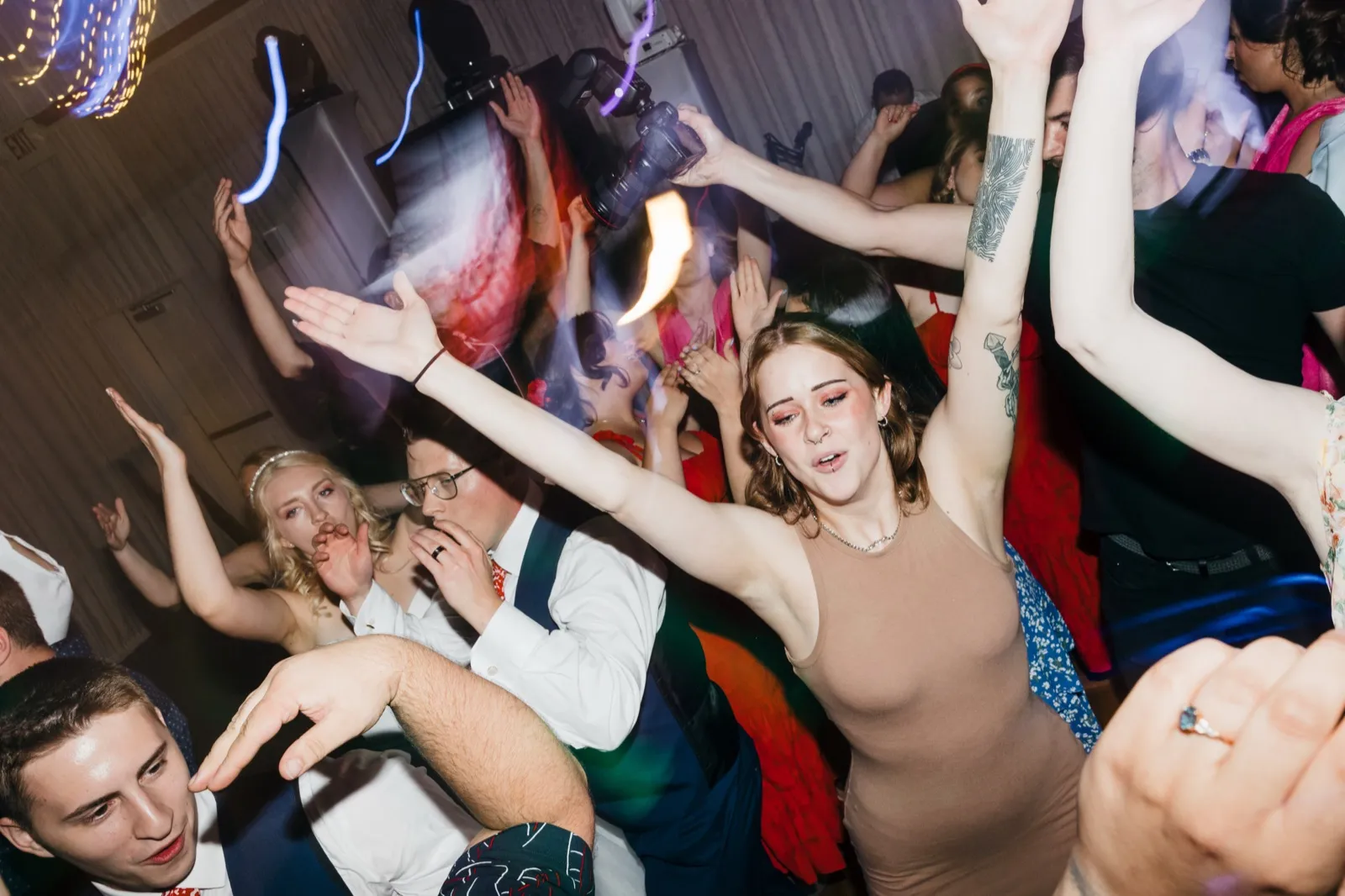 Guests pack the dance floor with arms raised as the bride cheers and the groom kisses her hand amid colored lights and motion blur. — Tim Larsen Photography, Brainerd Lakes MN