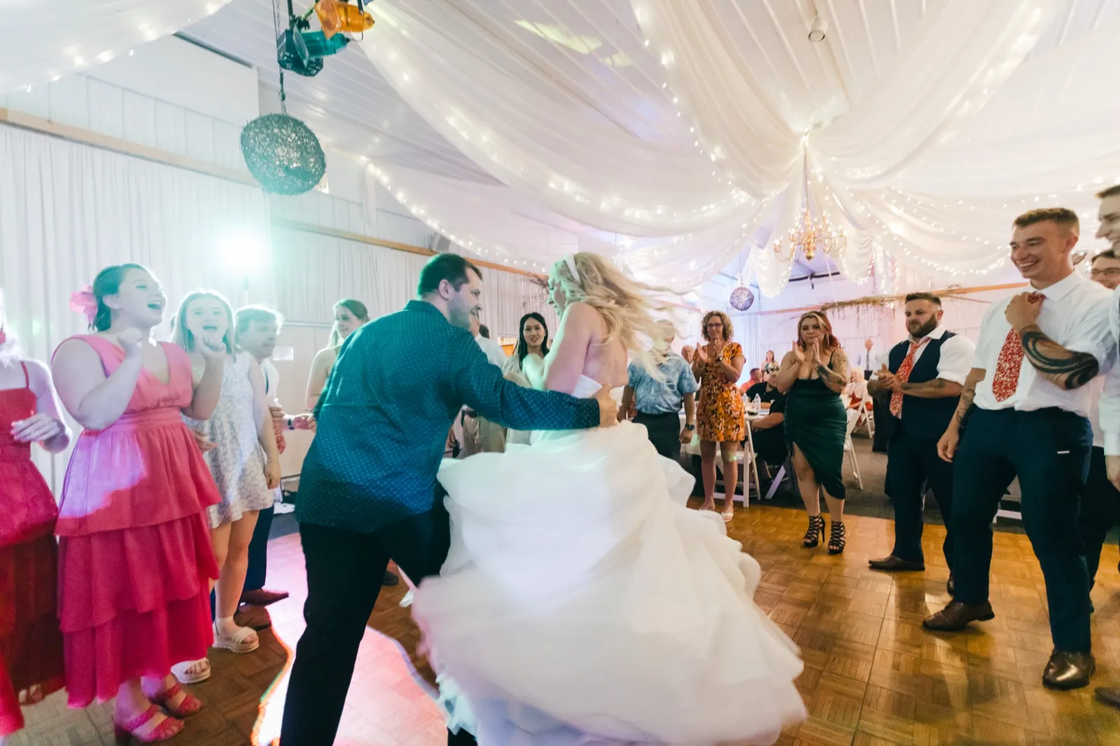 The groom spins the bride on the dance floor as her gown flares outward, surrounded by laughing bridal party and guests beneath draped fabric and string lights. — Tim Larsen Photography, Brainerd Lakes MN