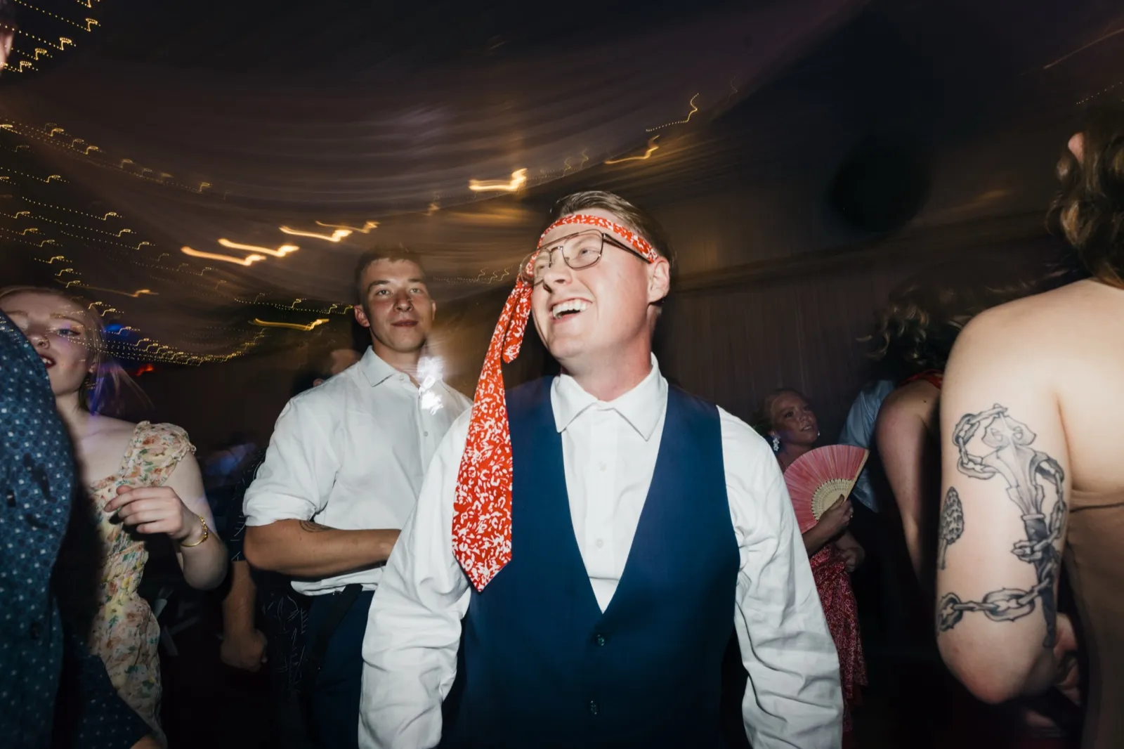 The groom laughs with his tie wrapped around his forehead like a headband as guests dance around him under a tented ceiling strung with lights. — Tim Larsen Photography, Brainerd Lakes MN