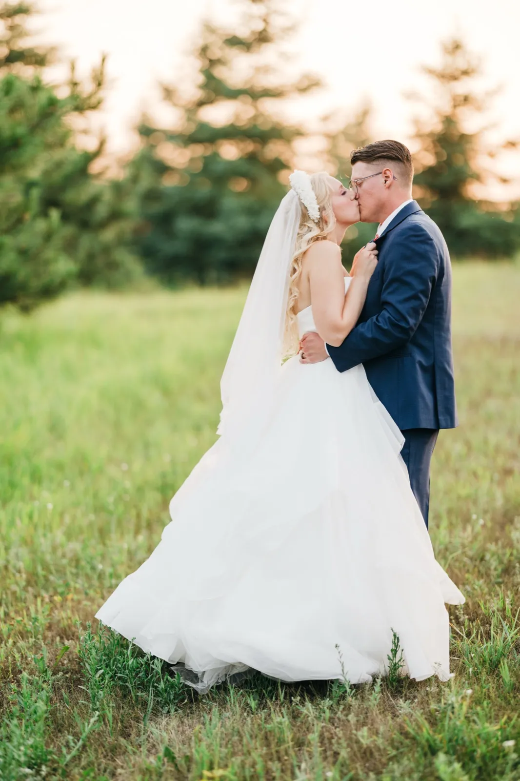 The bride and groom kiss in a sunlit meadow surrounded by evergreens, her veil and full gown trailing across the grass.