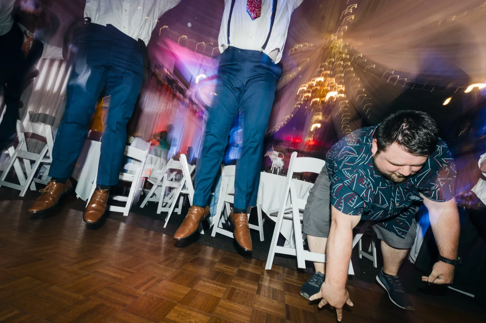 Three groomsmen stand on chairs dancing while a guest in a Hawaiian shirt crouches low on the dance floor under swirling string lights. — Tim Larsen Photography, Brainerd Lakes MN