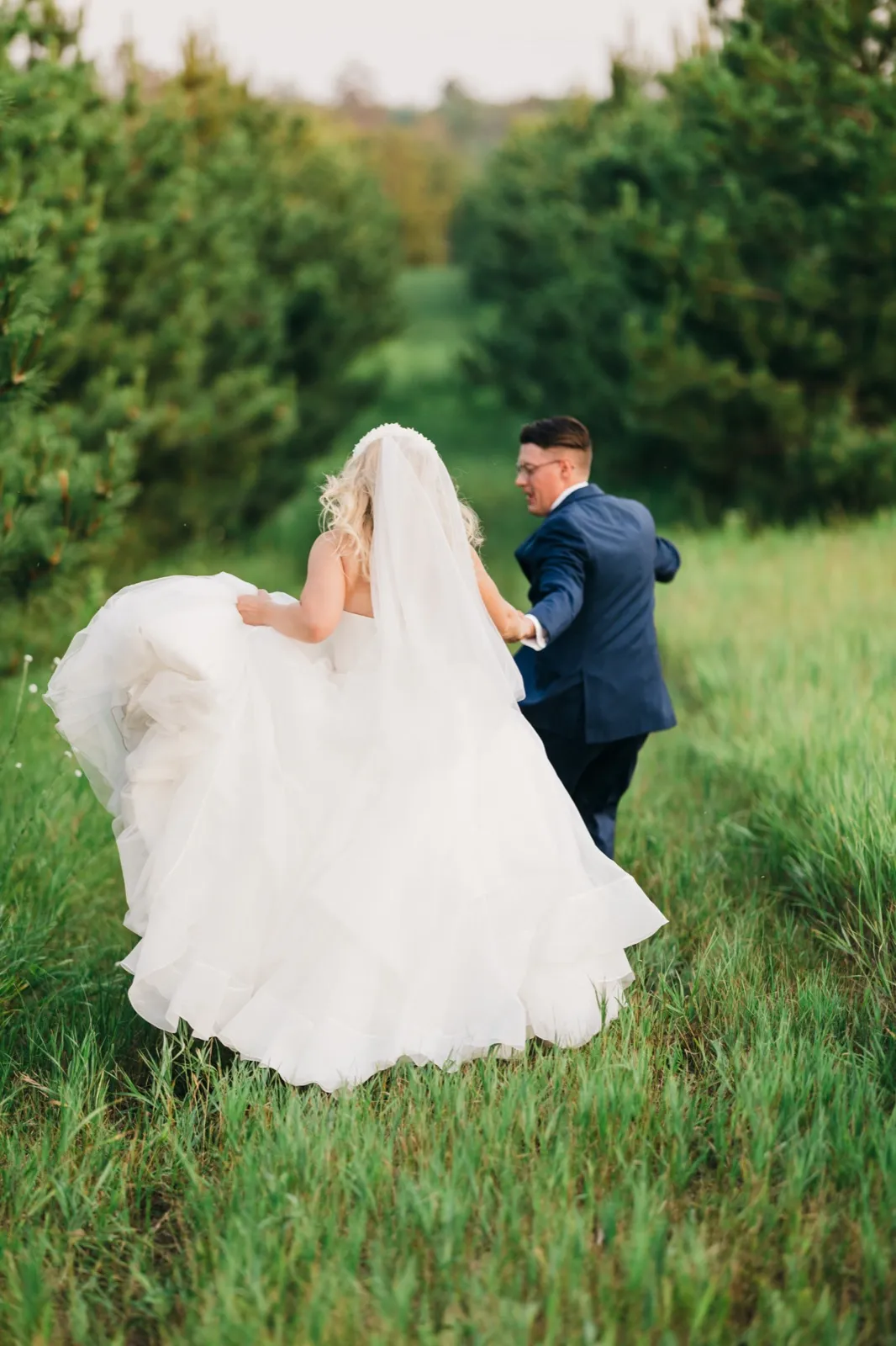 The bride lifts the skirt of her gown as the groom leads her by the hand down a corridor of tall pine trees. — Tim Larsen Photography, Brainerd Lakes MN