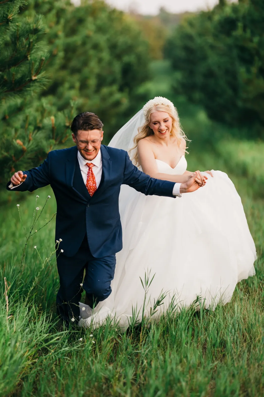 The bride and groom hold hands and run laughing through a row of evergreen trees, the bride lifting her white gown as her veil trails behind her. — Tim Larsen Photography, Brainerd Lakes MN