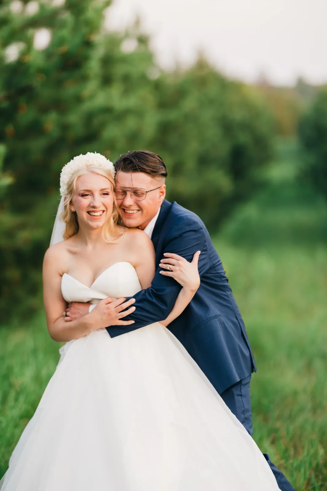 The groom embraces the bride from behind as both laugh in a grassy field with evergreens behind them. — Tim Larsen Photography, Brainerd Lakes MN