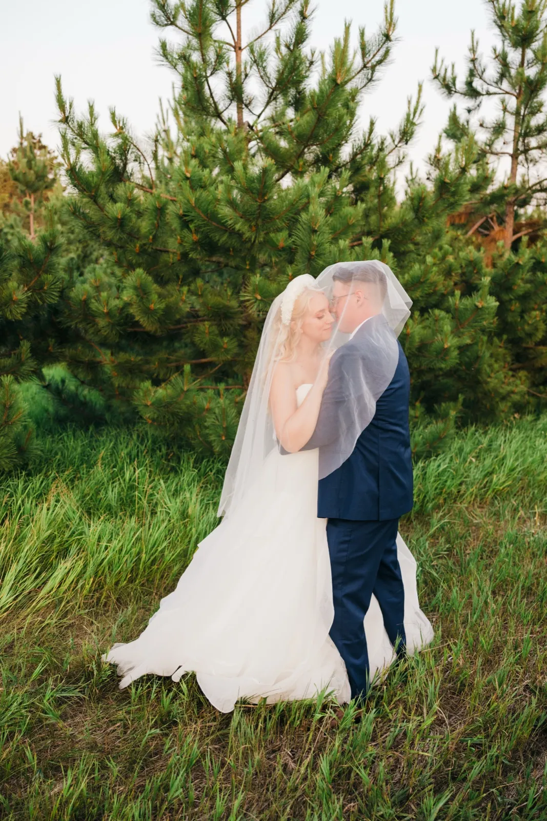 The bride and groom stand forehead-to-forehead beneath her draped veil in a clearing surrounded by tall pine trees. — Tim Larsen Photography, Brainerd Lakes MN