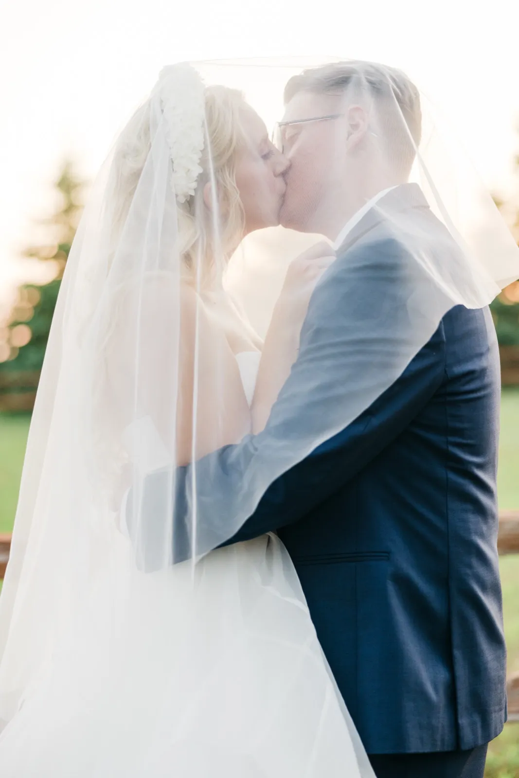 The bride and groom kiss while her long veil drapes over both of them, backlit by golden evening light. — Tim Larsen Photography, Brainerd Lakes MN