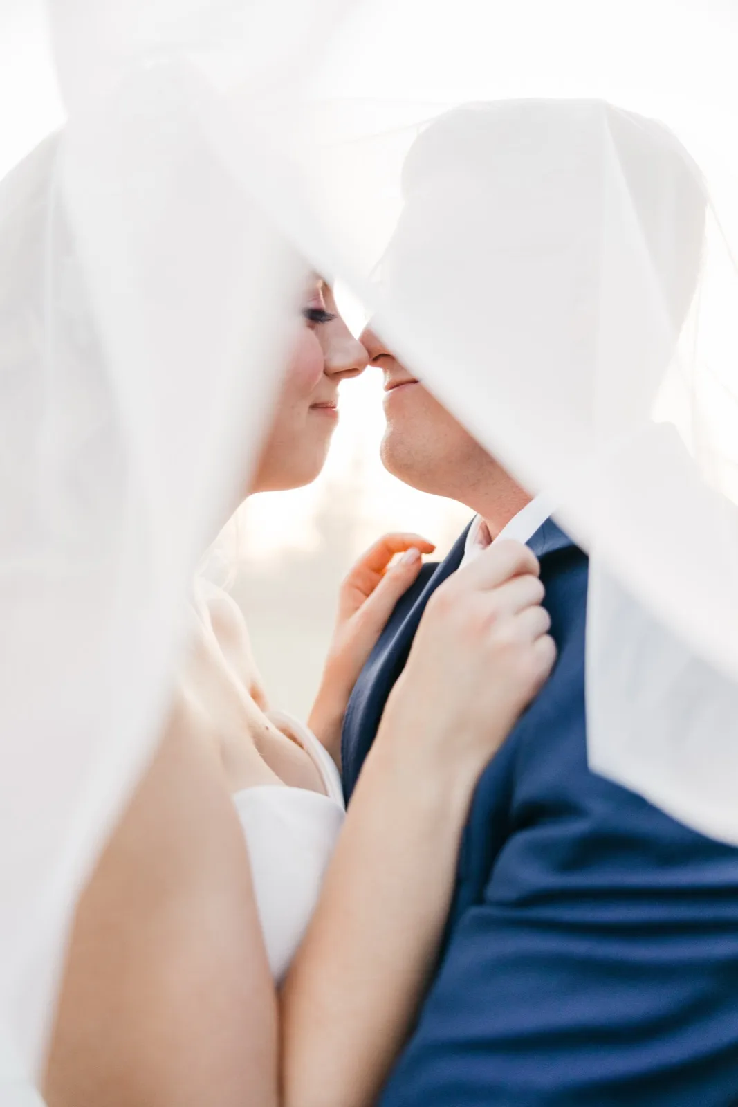 The bride and groom lean in nose-to-nose beneath her veil, which drapes around them and glows in the backlight as she holds his lapel. — Tim Larsen Photography, Brainerd Lakes MN
