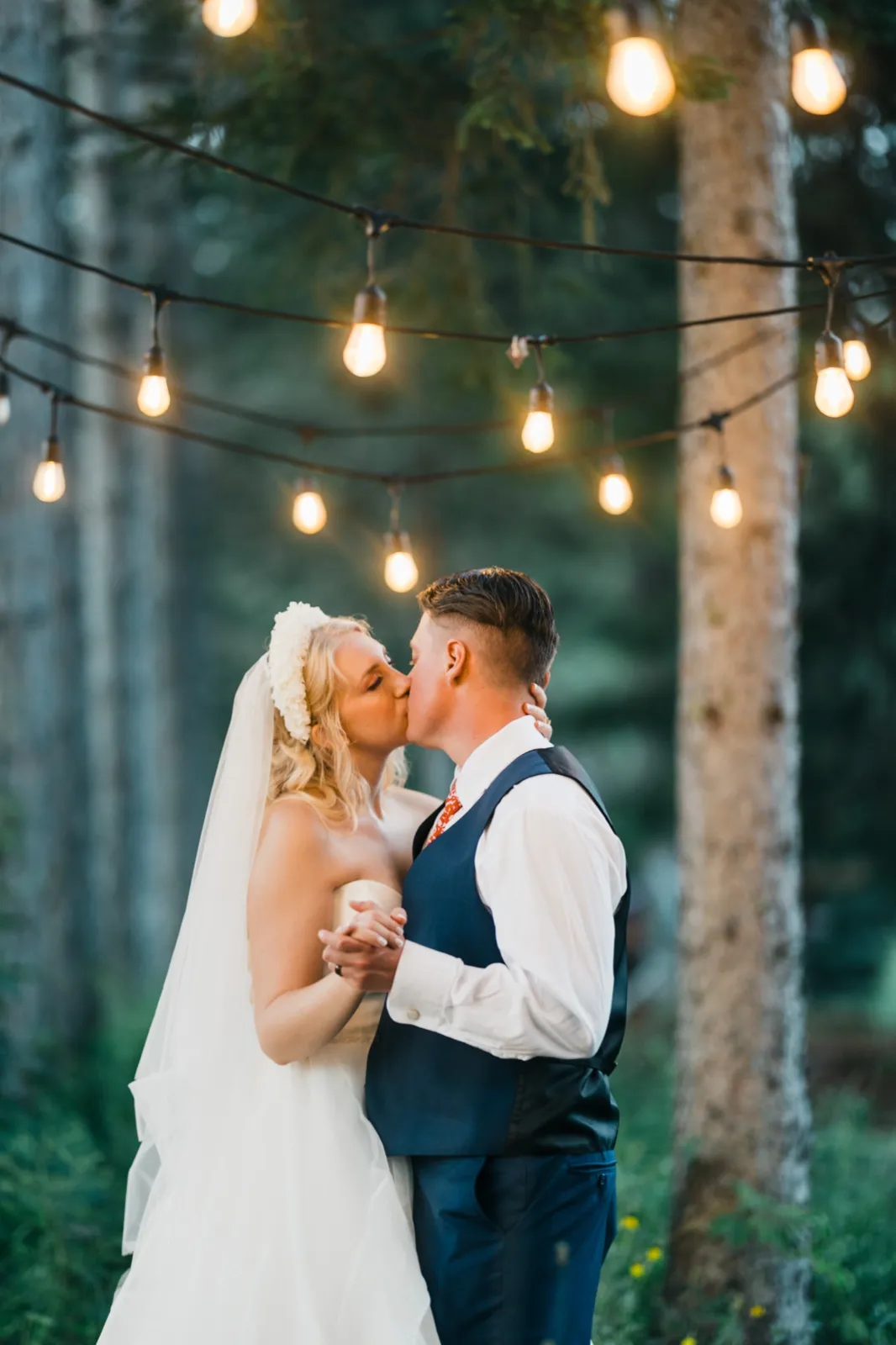 The couple kisses in a forest clearing beneath strands of glowing Edison-bulb string lights strung between tall pine trees at dusk. — Tim Larsen Photography, Brainerd Lakes MN