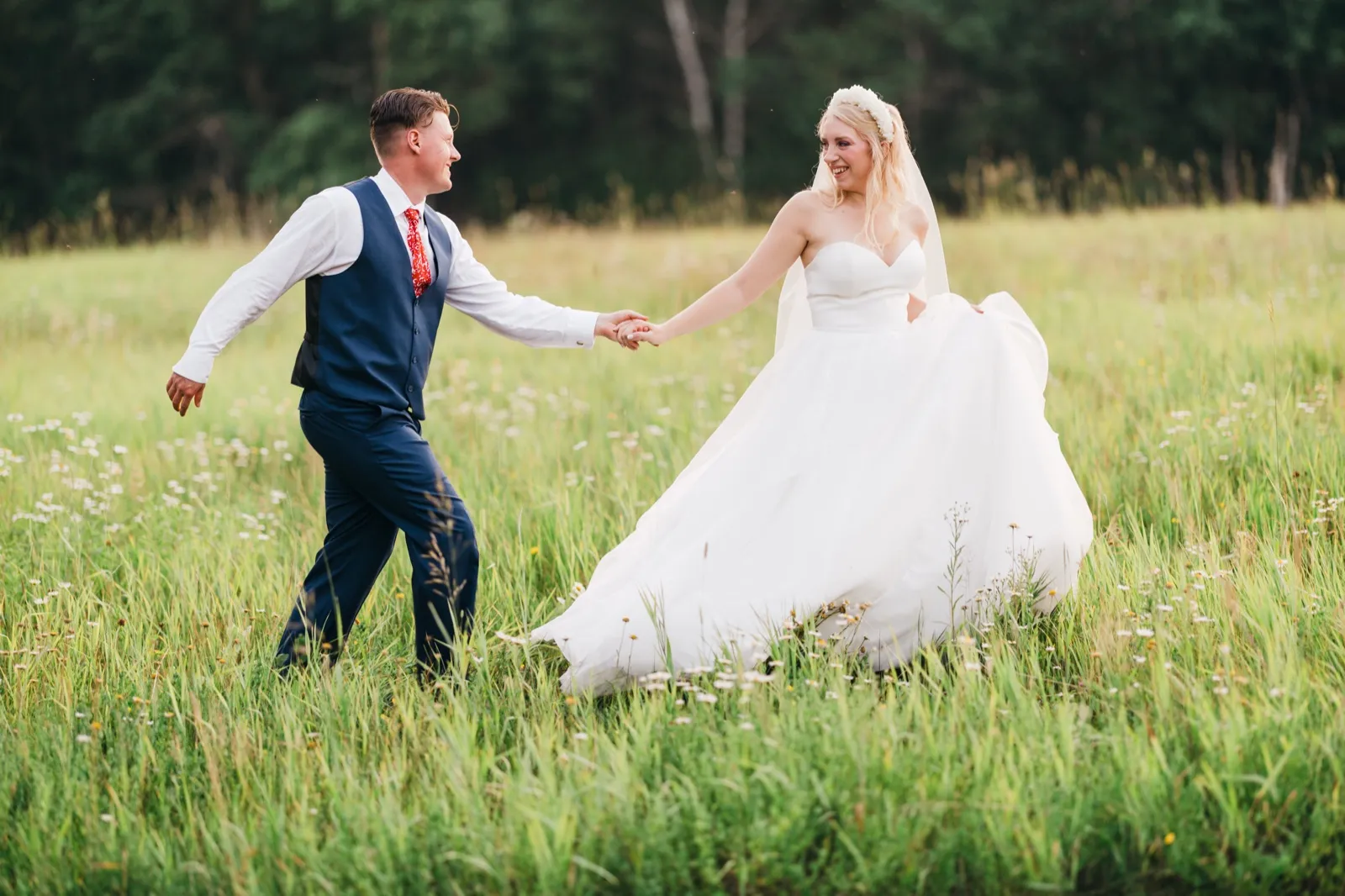 The groom leads the bride by the hand through a tall wildflower meadow, her gown trailing behind her as they both smile. — Tim Larsen Photography, Brainerd Lakes MN