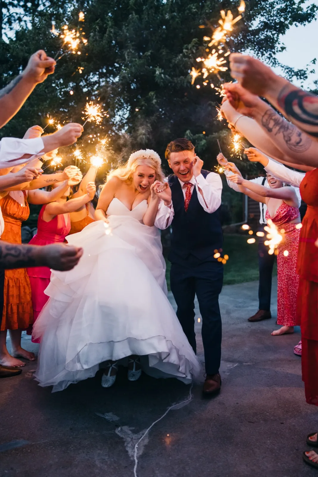 The bride and groom laugh wide-mouthed as they walk through a tunnel of guests holding sparklers at dusk.