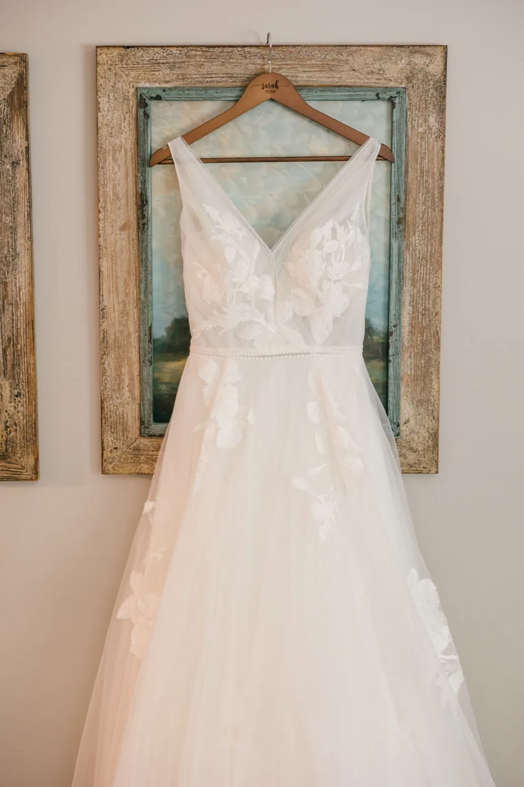 A lace and tulle wedding dress hangs from a personalized wooden hanger reading Sarah, centered against a weathered turquoise framed painting on a neutral wall — Tim Larsen Photography, Brainerd Lakes MN