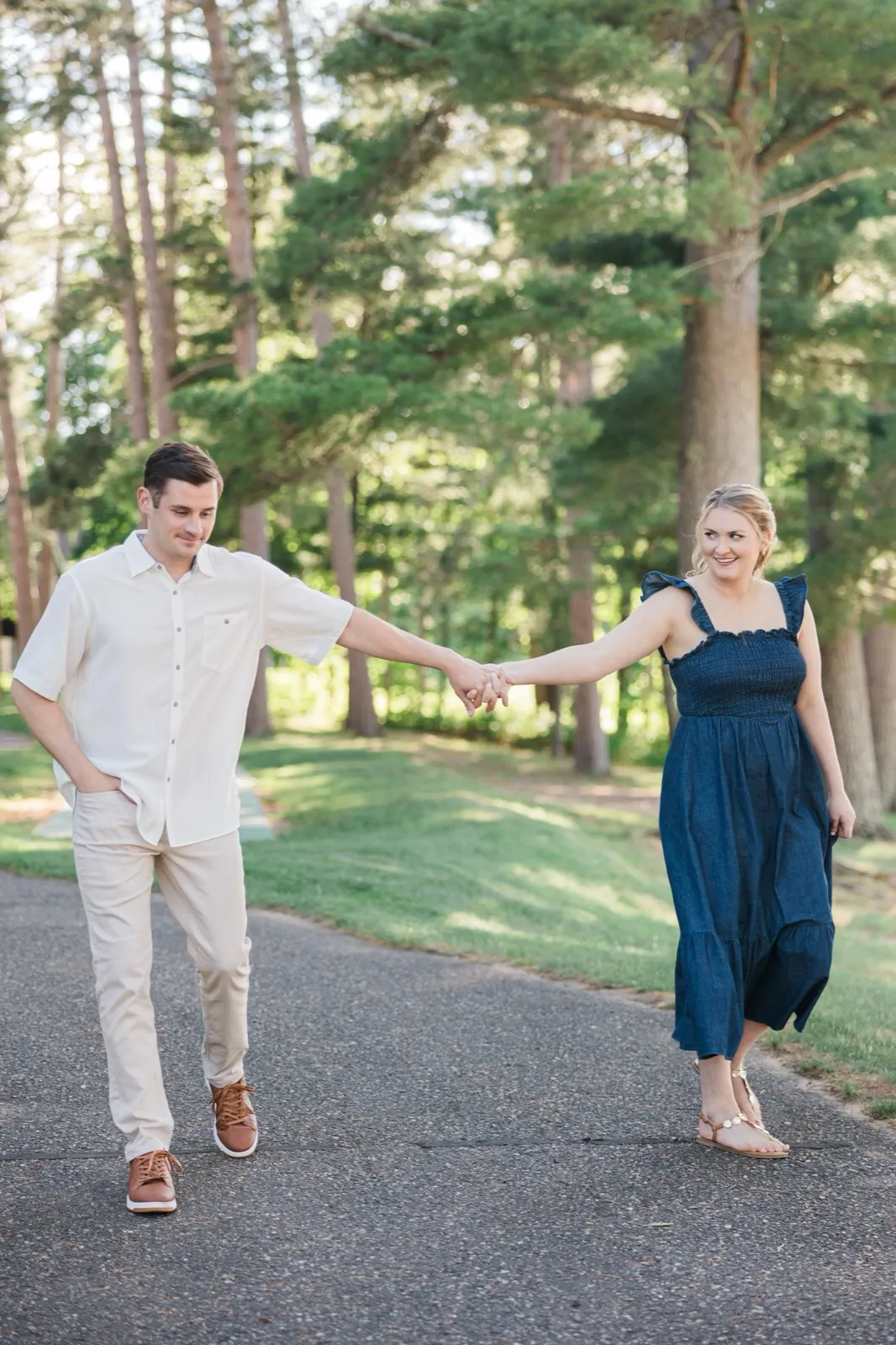 An engaged couple walk hand in hand down a pine-lined drive on the Roy Lake side of Grand View Lodge in evening light — Tim Larsen Photography