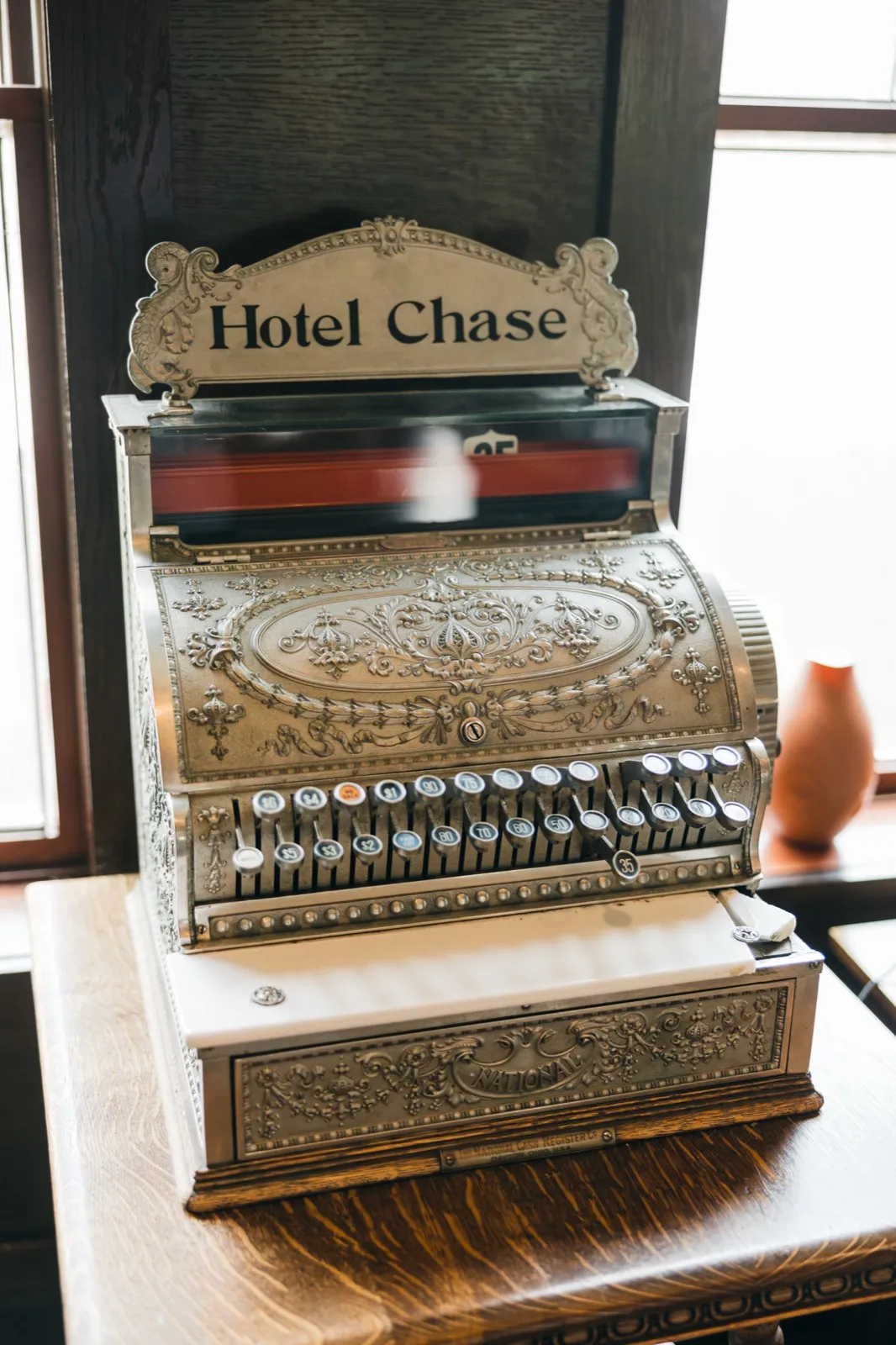 An antique ornate silver National cash register with a 'Hotel Chase' nameplate sits on a dark wood counter near bright windows. — Tim Larsen Photography, Brainerd Lakes MN