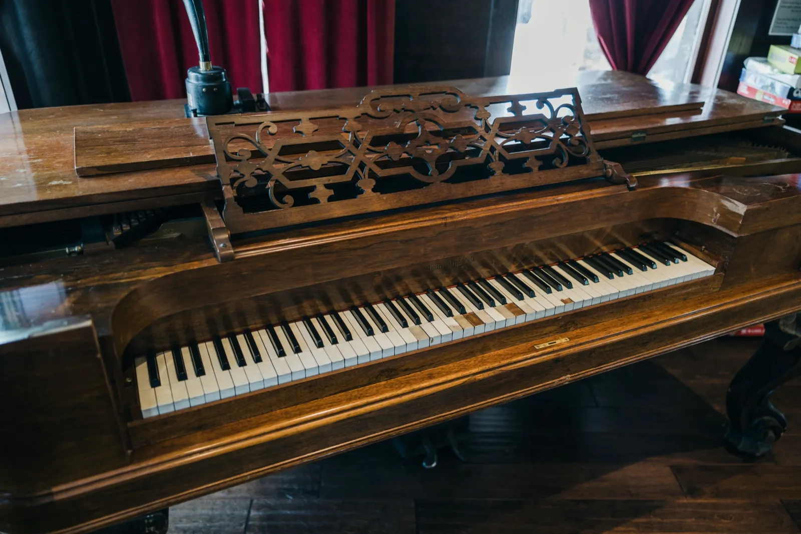 An antique wooden square grand piano with an ornate carved music rack sits in a window-lit parlor with deep red curtains. — Tim Larsen Photography, Brainerd Lakes MN