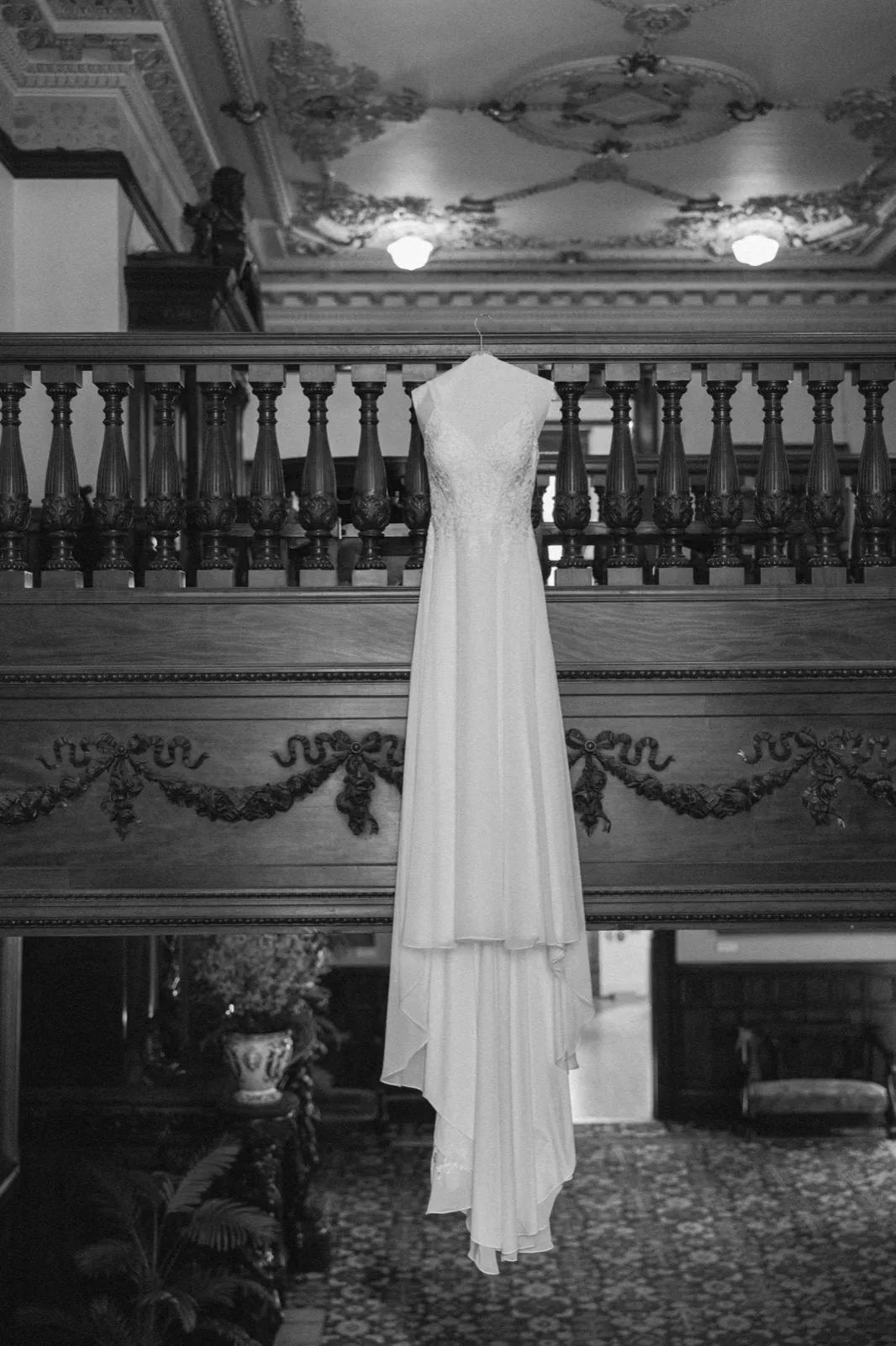 A wedding dress hangs from the ornate carved wooden balustrade of a grand staircase, its train cascading down over the dark paneled wall in a black-and-white image of the American Swedish Institute interior. — Tim Larsen Photography, Minneapolis MN