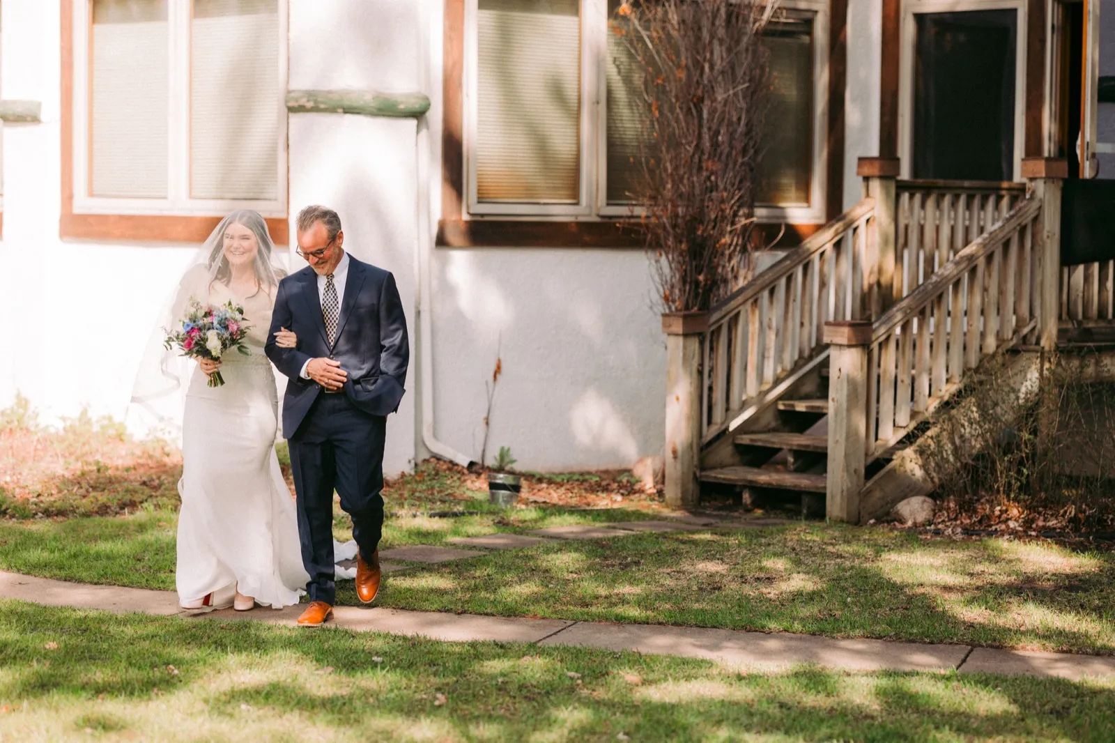 The bride, smiling broadly behind her veil and holding a colorful bouquet, walks down a stone path with her father along the side of the lodge — Tim Larsen Photography, Brainerd Lakes MN