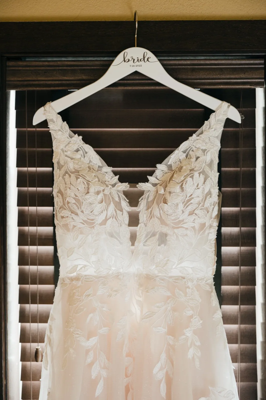 A lace wedding dress hangs on a personalized 'bride' hanger dated 7-16-2022 in front of a shuttered window. — Tim Larsen Photography, Brainerd Lakes MN