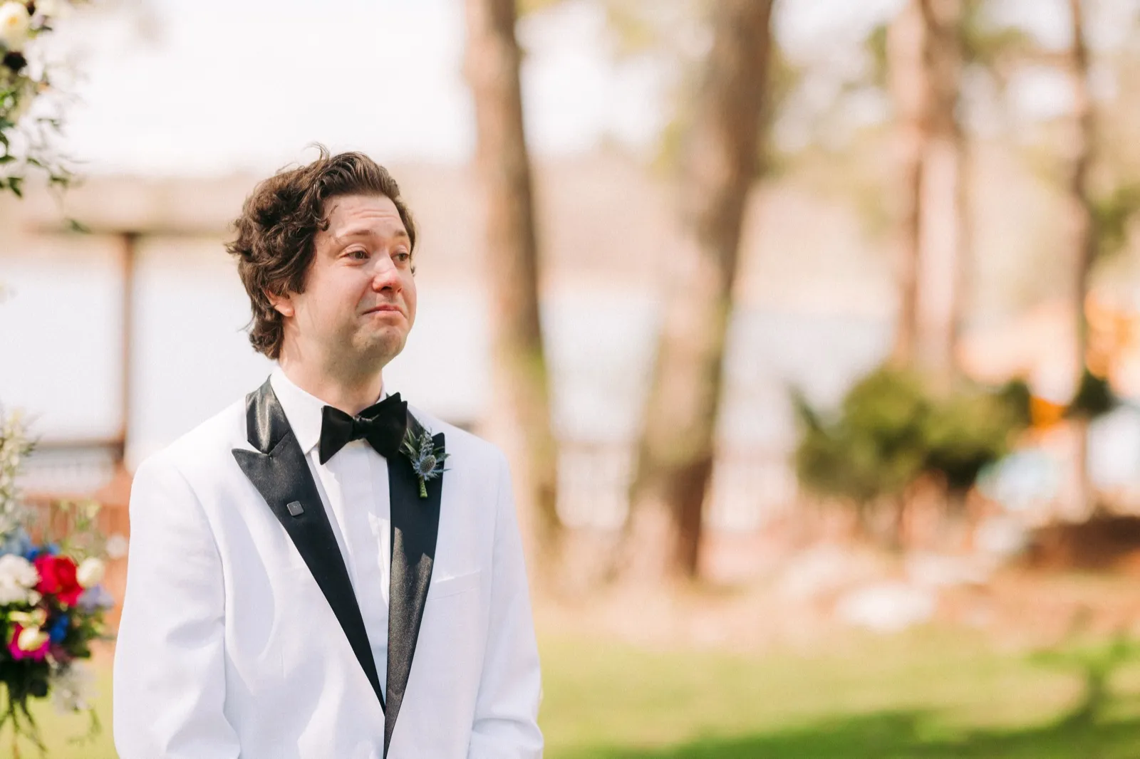 The groom in a white tuxedo jacket fights back tears at the ceremony altar as he watches his bride approach down the aisle at the lakeside ceremony — Tim Larsen Photography, Brainerd Lakes MN