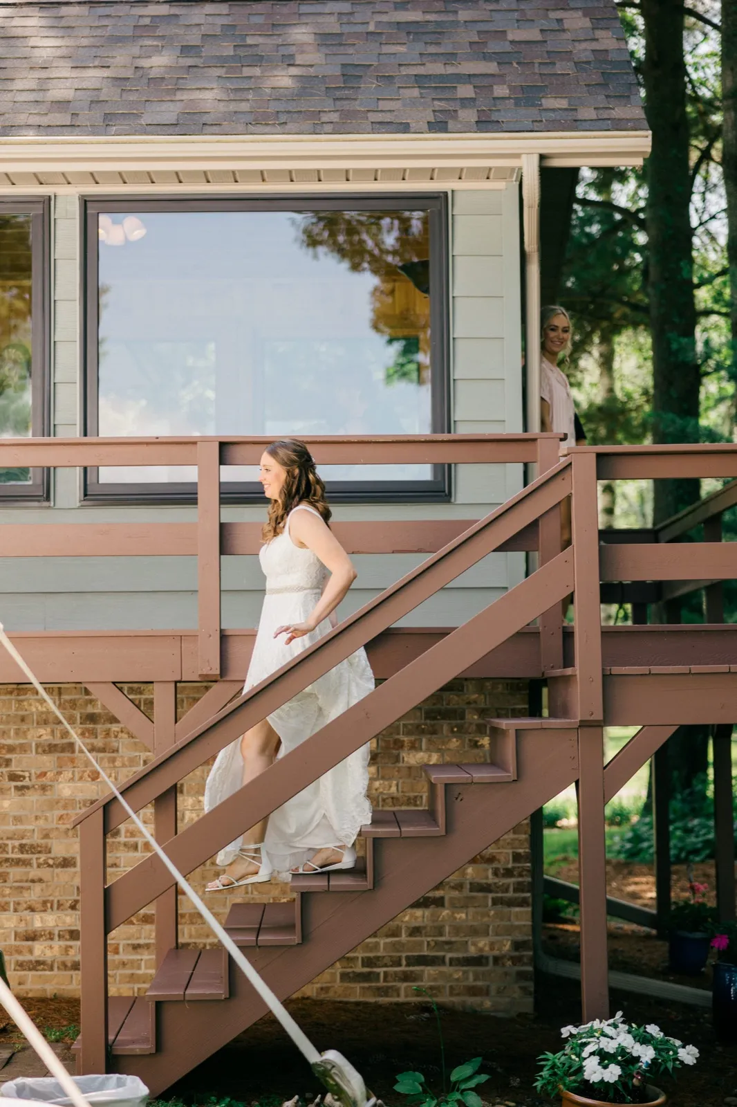 The bride lifts her gown as she walks down a wooden cabin deck staircase while a bridesmaid watches from the doorway above. — Tim Larsen Photography, Brainerd Lakes MN