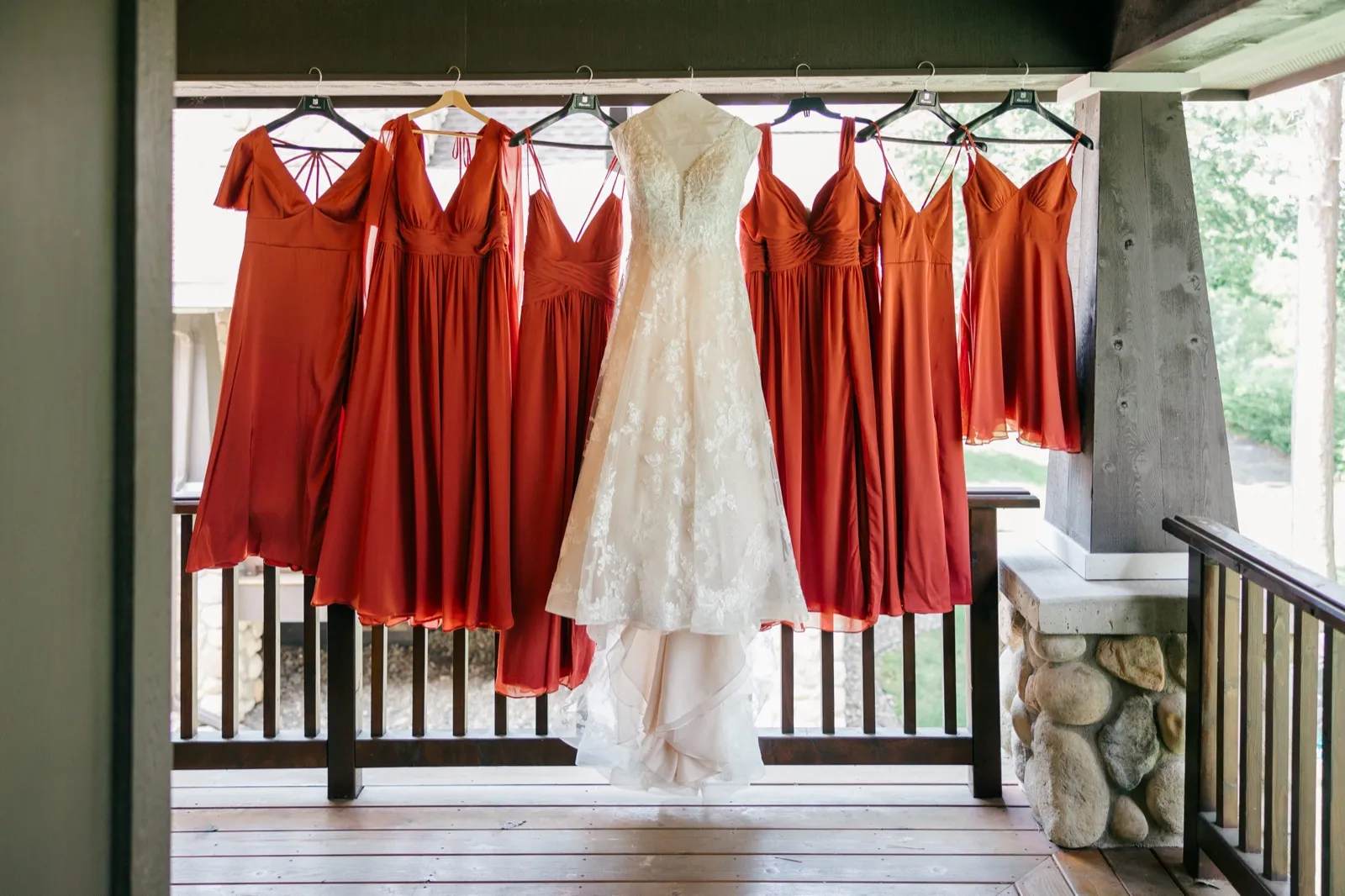 A wedding gown hangs at the center of six rust-colored bridesmaid dresses suspended from a wooden porch railing with a stone fireplace visible to the right — Tim Larsen Photography, Brainerd Lakes MN