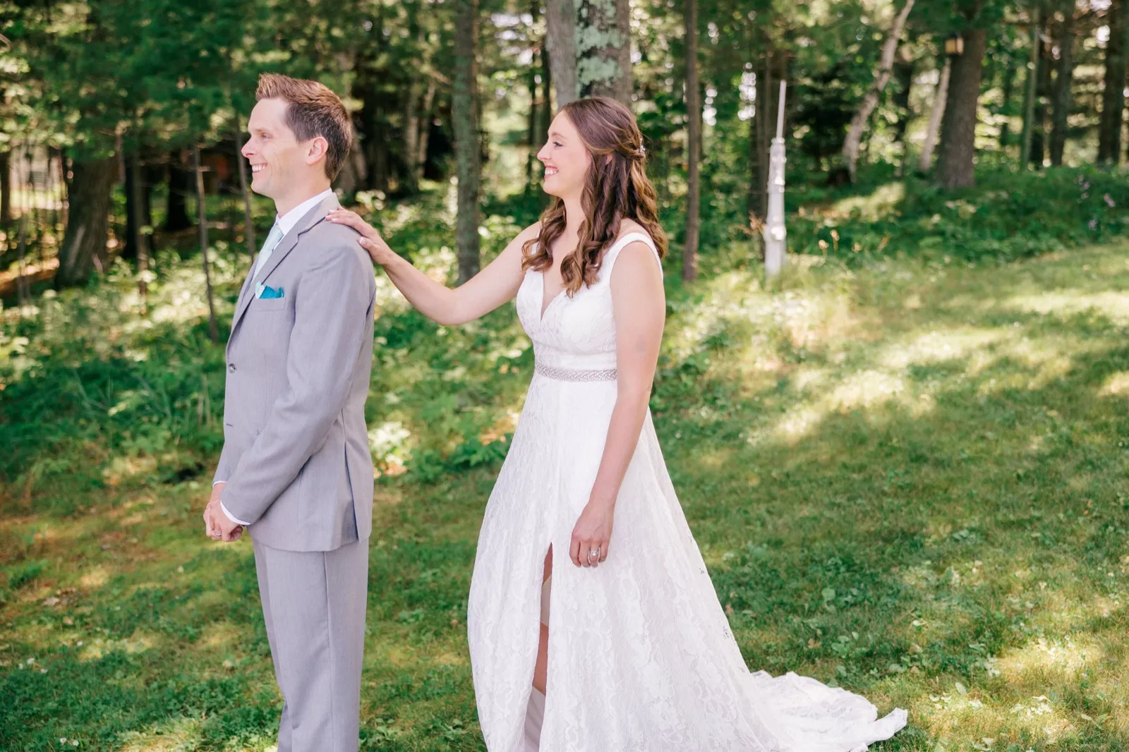 The bride reaches her hand to the groom's shoulder from behind in a sunlit wooded clearing just before he turns for their first look. — Tim Larsen Photography, Brainerd Lakes MN