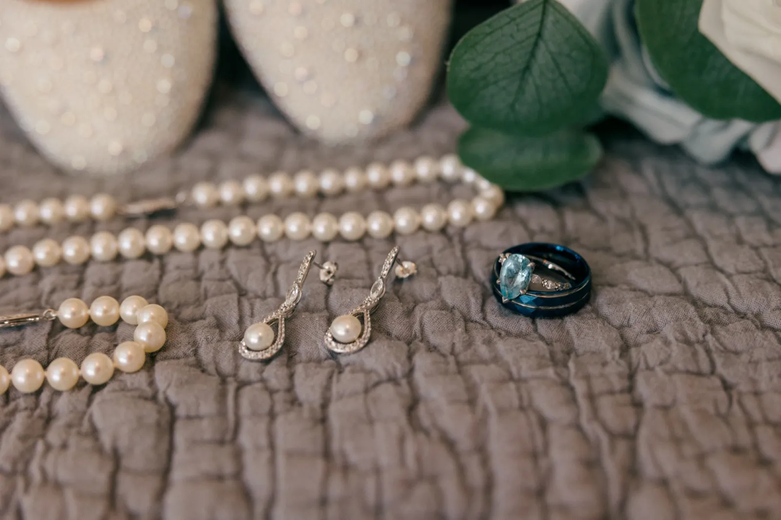 A pearl necklace, pearl drop earrings, and a pear-cut aquamarine engagement ring with a dark wedding band rest on a textured gray linen near sparkling bridal shoes and eucalyptus — Tim Larsen Photography, Brainerd Lakes MN