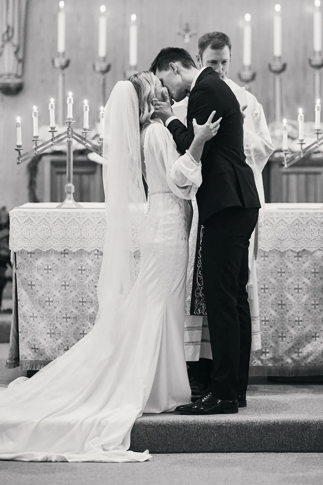 A bride and groom share their first kiss at a candlelit church altar while the officiant stands behind them, the bride's long cathedral veil trailing across the steps. — Tim Larsen Photography, Brainerd Lakes MN
