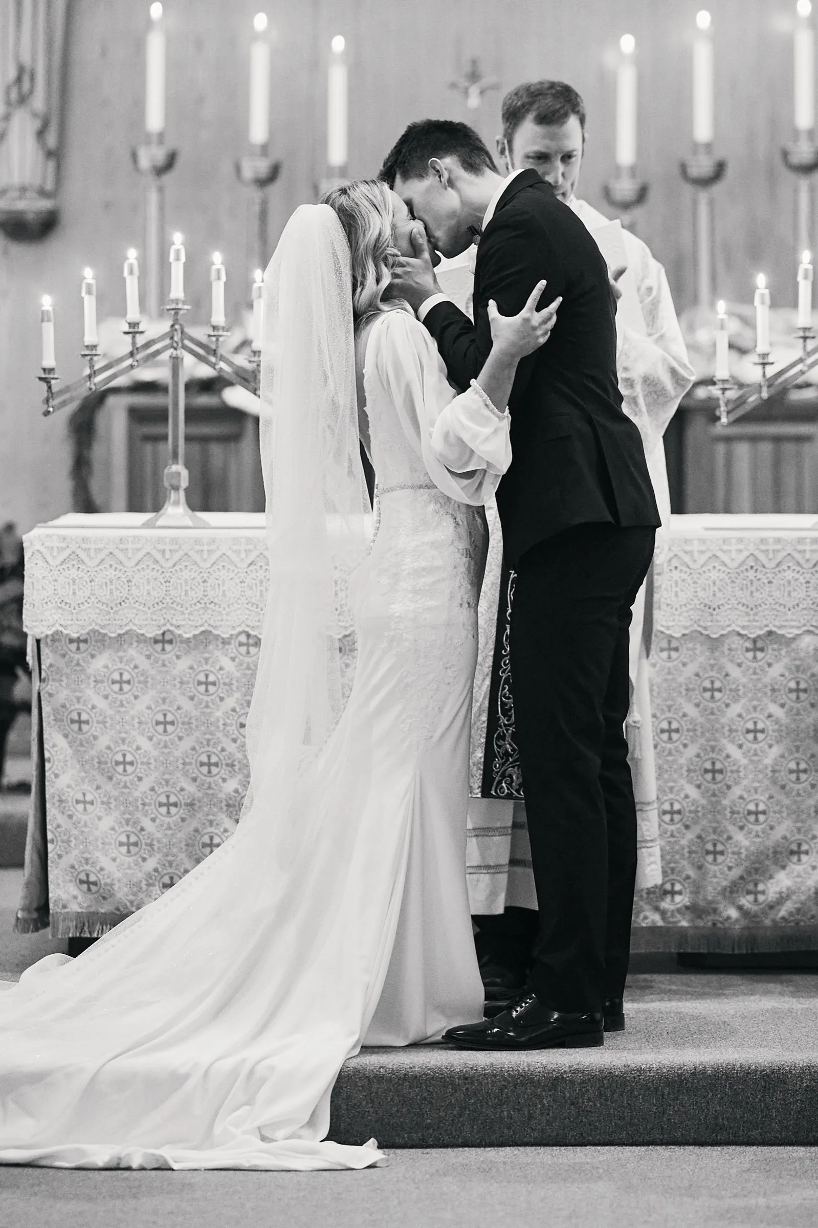 Madeline and Sam share their first kiss at a candlelit church altar while the priest stands behind them, her cathedral veil trailing across the steps — Tim Larsen Photography, Brainerd Lakes MN