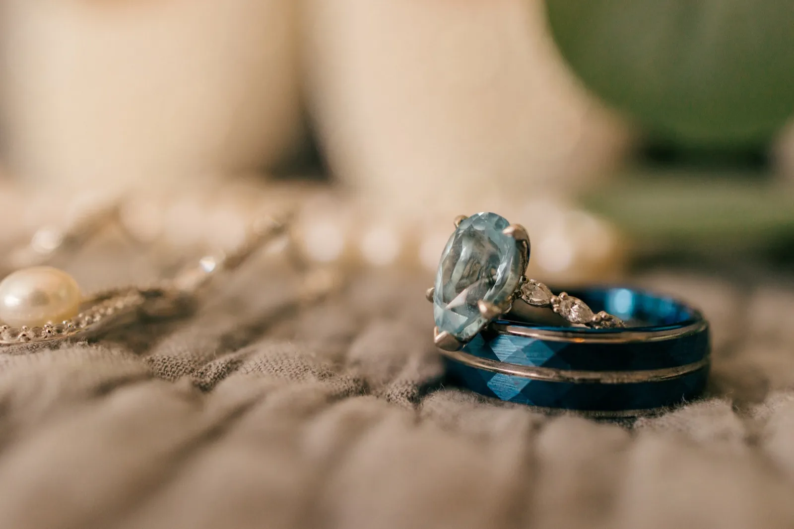 A pear-shaped aquamarine engagement ring rests atop a blue tungsten wedding band on soft fabric, with pearls and a delicate chain blurred in the foreground — Tim Larsen Photography, Brainerd Lakes MN