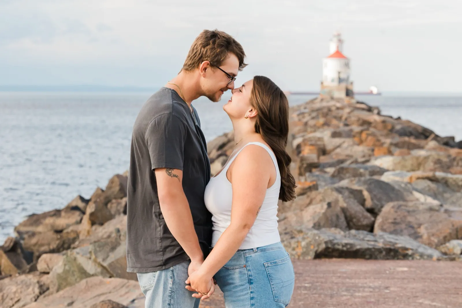 Reeden and Jacklyn stand nose to nose on the rocky Wisconsin Point breakwater, holding hands, the Superior Entry lighthouse and a passing freighter behind them — Tim Larsen Photography