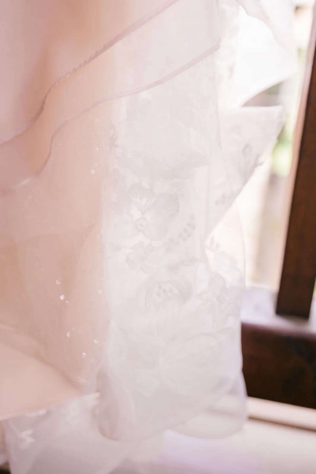 Backlit close-up of a lace and tulle wedding veil hung in a window, the floral embroidery glowing translucent in soft morning light — Tim Larsen Photography, Brainerd Lakes MN