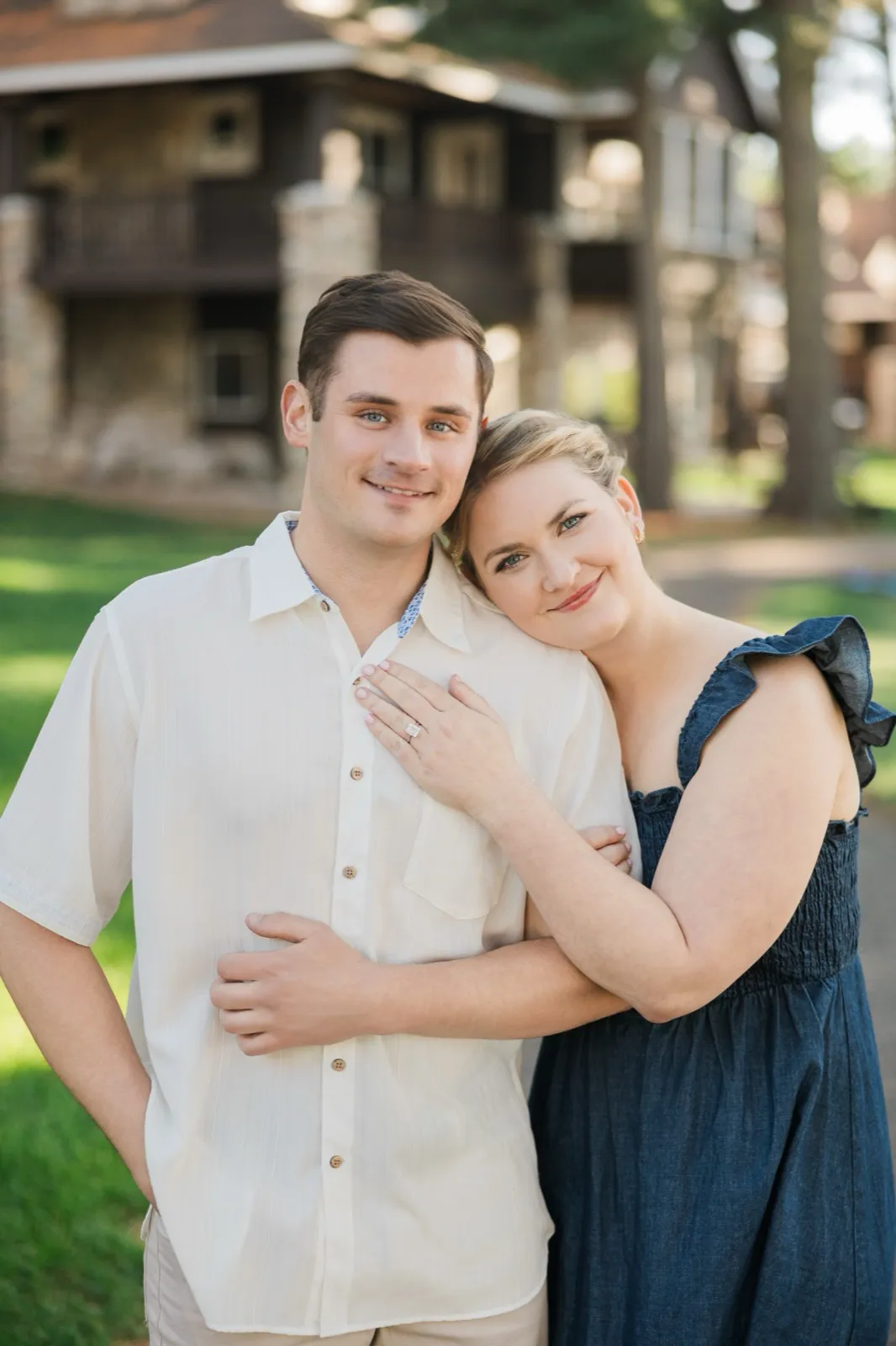 An engaged couple pose in front of the rustic log cabins on the Roy Lake side of Grand View Lodge, the woman's ring hand on her partner's chest — Tim Larsen Photography