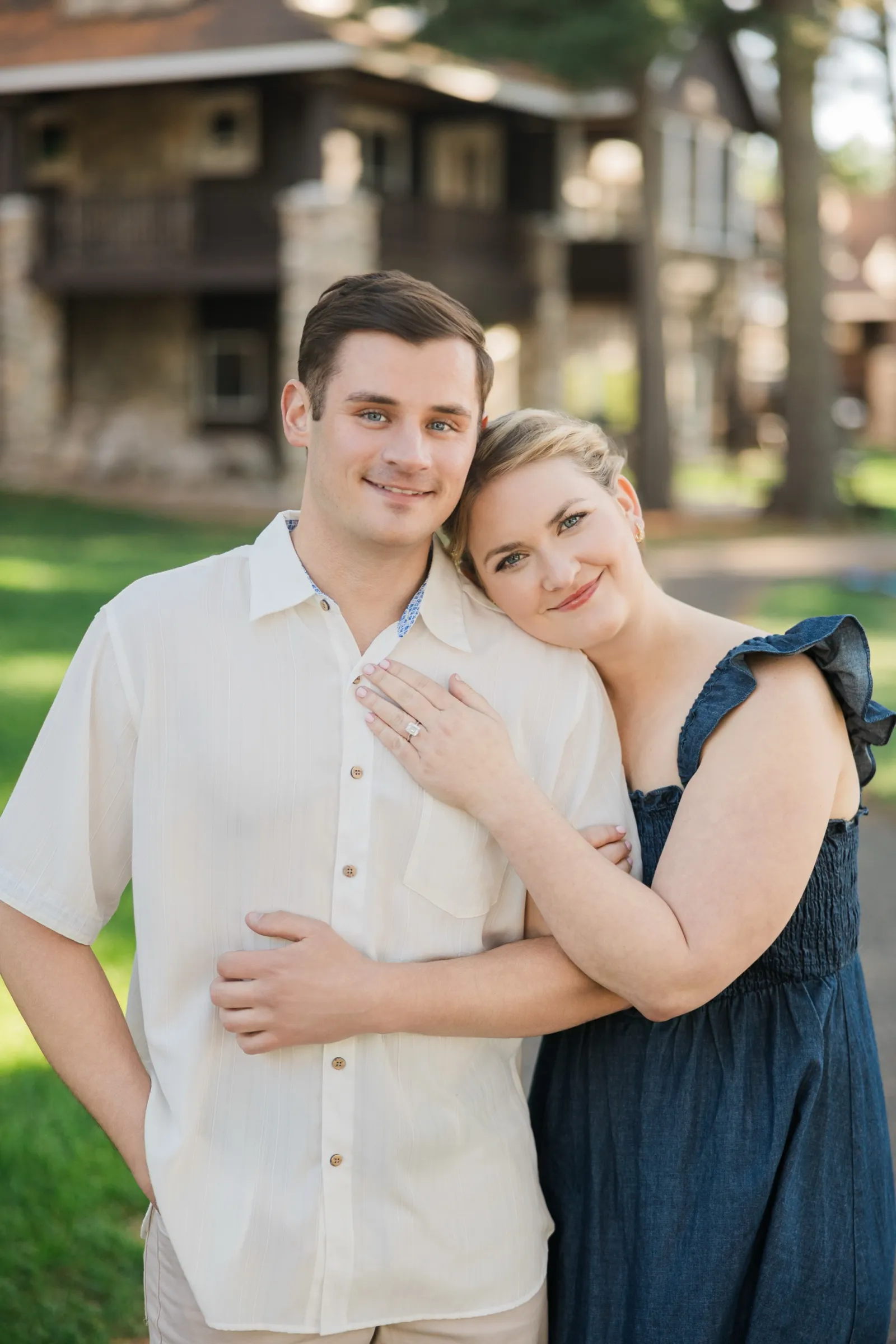 An engaged couple pose in front of the rustic log cabins on the Roy Lake side of Grand View Lodge, the woman's ring hand on her partner's chest — Tim Larsen Photography