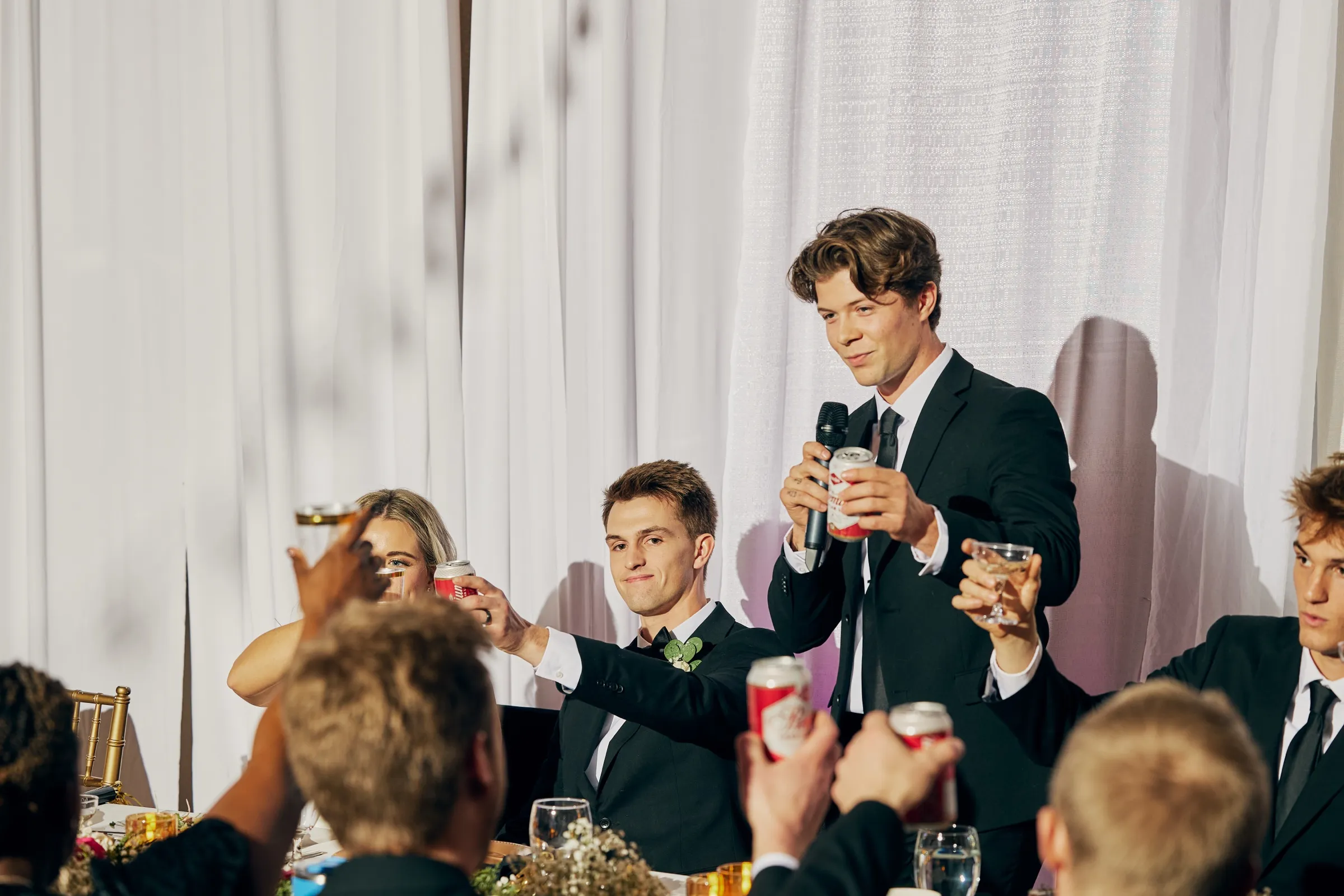 The best man holds a microphone and a beer can as Sam and surrounding guests raise drinks in a toast at the head table — Tim Larsen Photography, Brainerd Lakes MN