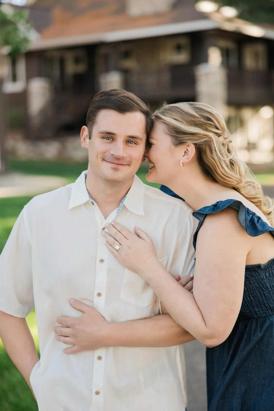 A woman whispers into her fiance's ear, her engagement ring hand on his chest, by the log cabins on Grand View Lodge's Roy Lake side — Tim Larsen Photography