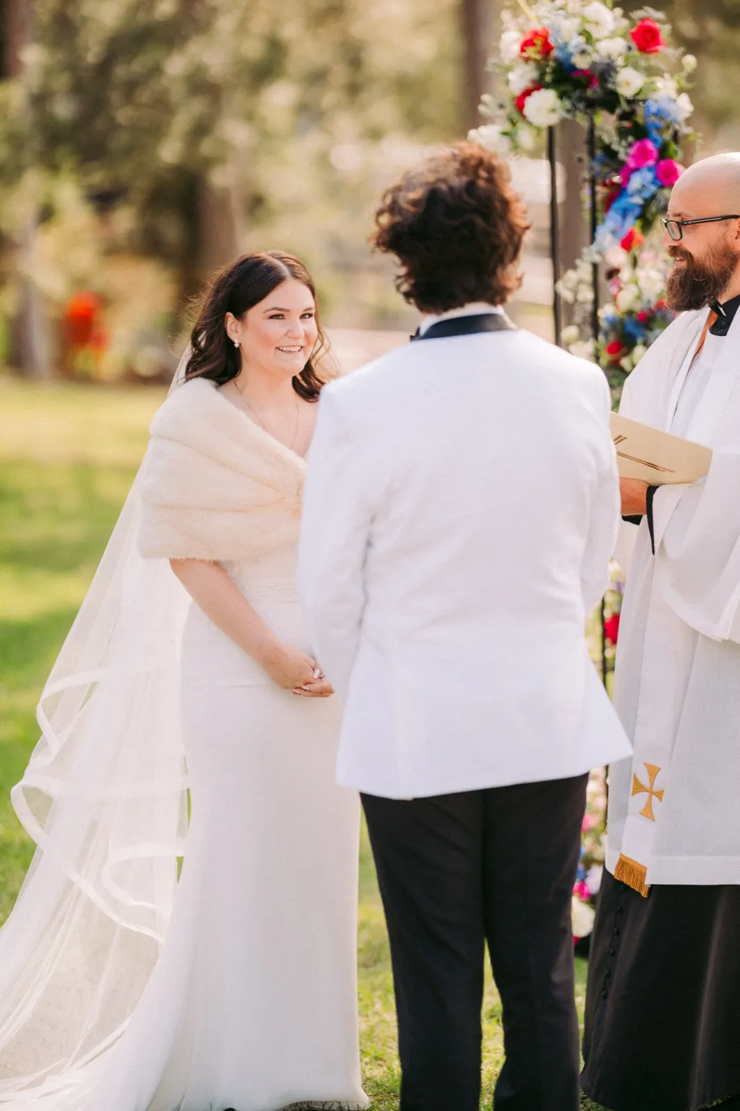 The bride smiles warmly at the groom during their outdoor ceremony as the officiant stands beside them in front of a colorful floral arch — Tim Larsen Photography, Brainerd Lakes MN