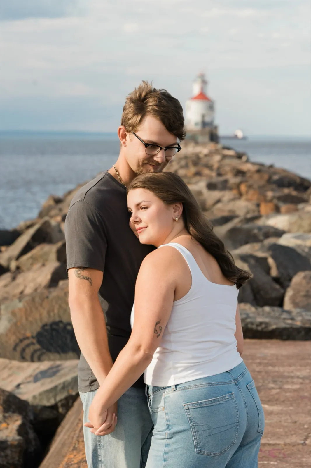 Jacklyn rests her head on Reeden's chest and looks toward the camera while he gazes down at her, the lighthouse soft behind them — Tim Larsen Photography