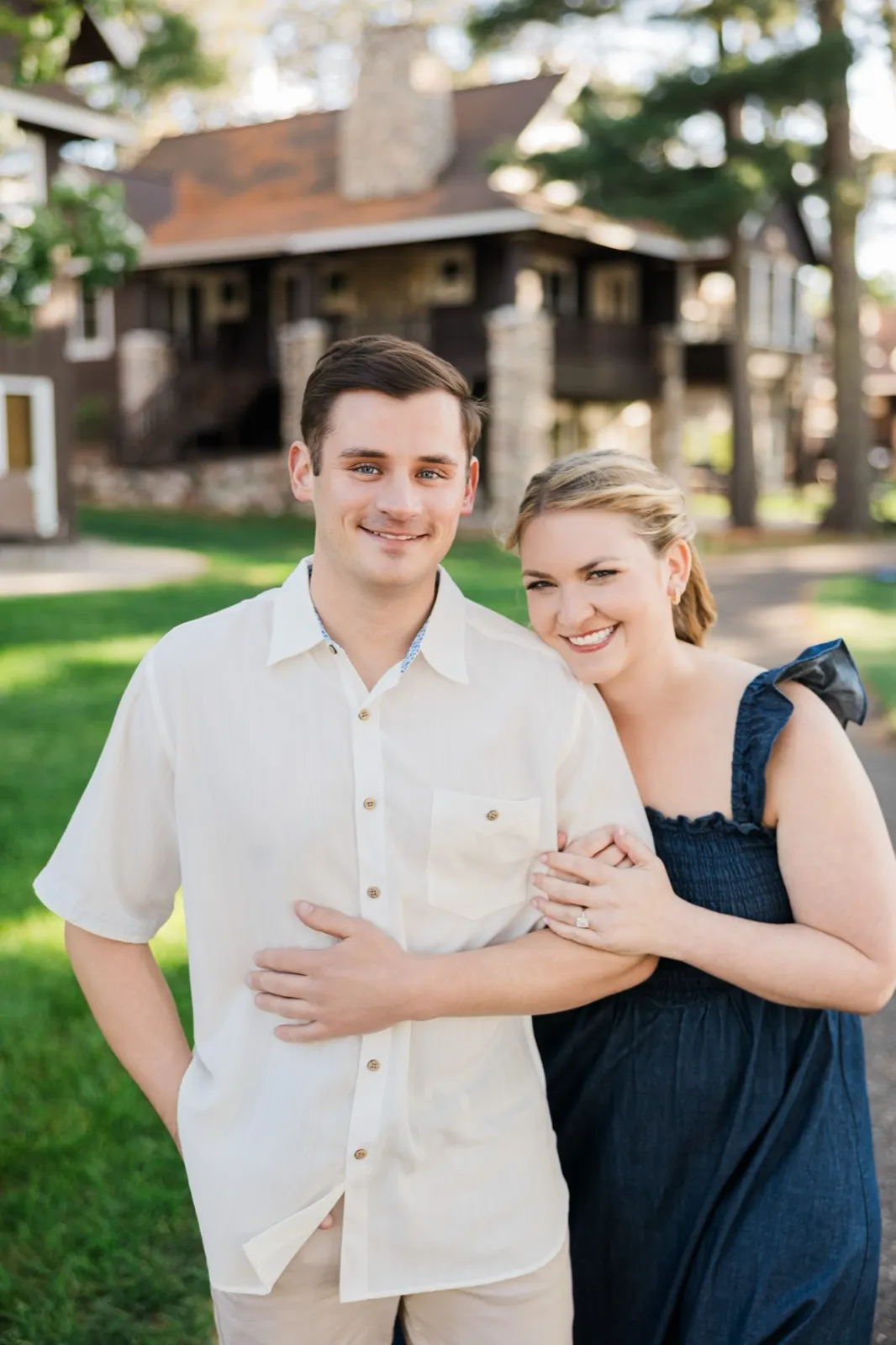 An engaged couple smile at the camera in front of the rustic log buildings on Grand View Lodge's Roy Lake side — Tim Larsen Photography