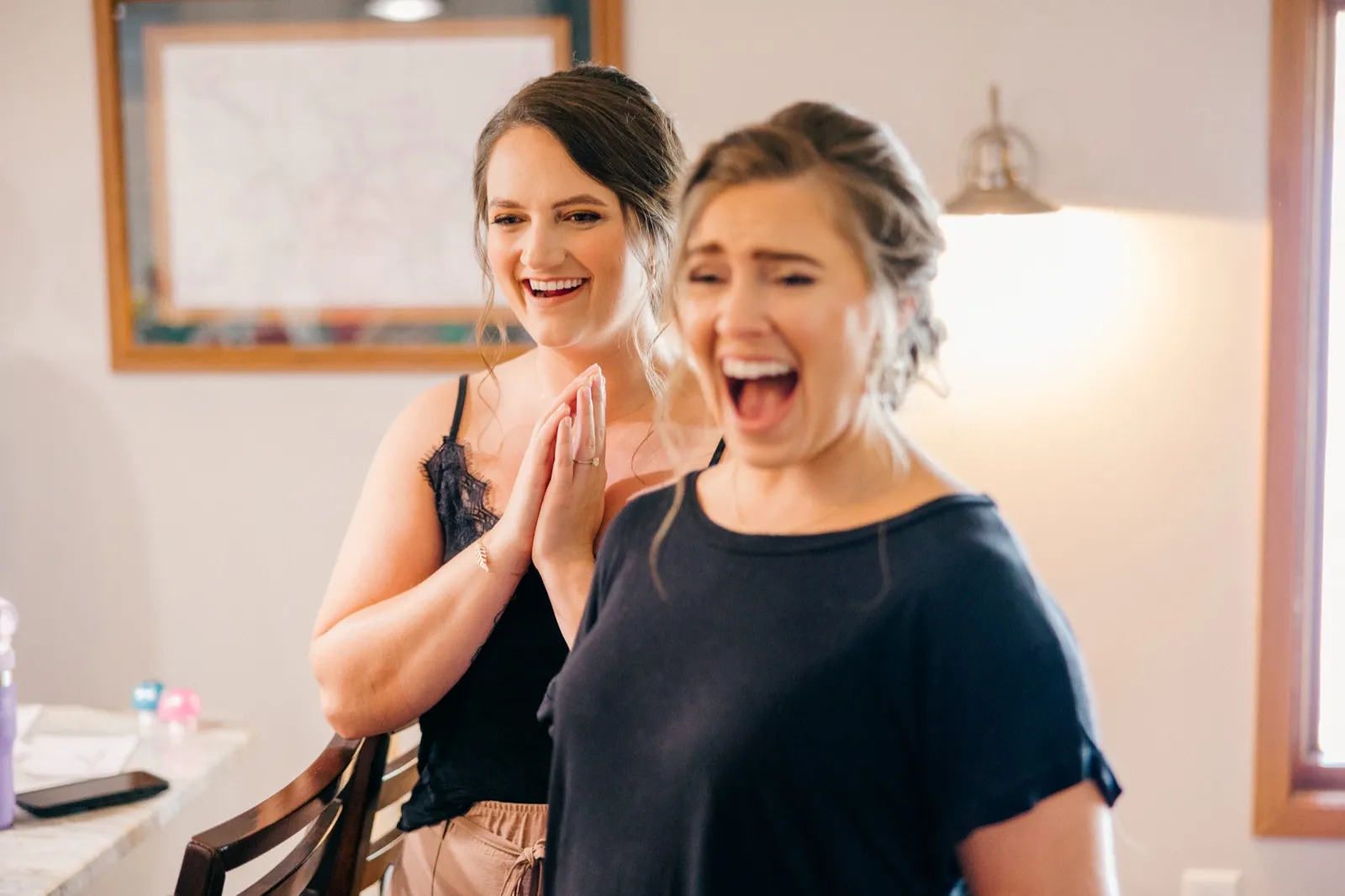 A bridesmaid presses her hands together and laughs while another woman in the foreground reacts with an open-mouthed laugh during getting-ready inside the cabin — Tim Larsen Photography, Brainerd Lakes MN