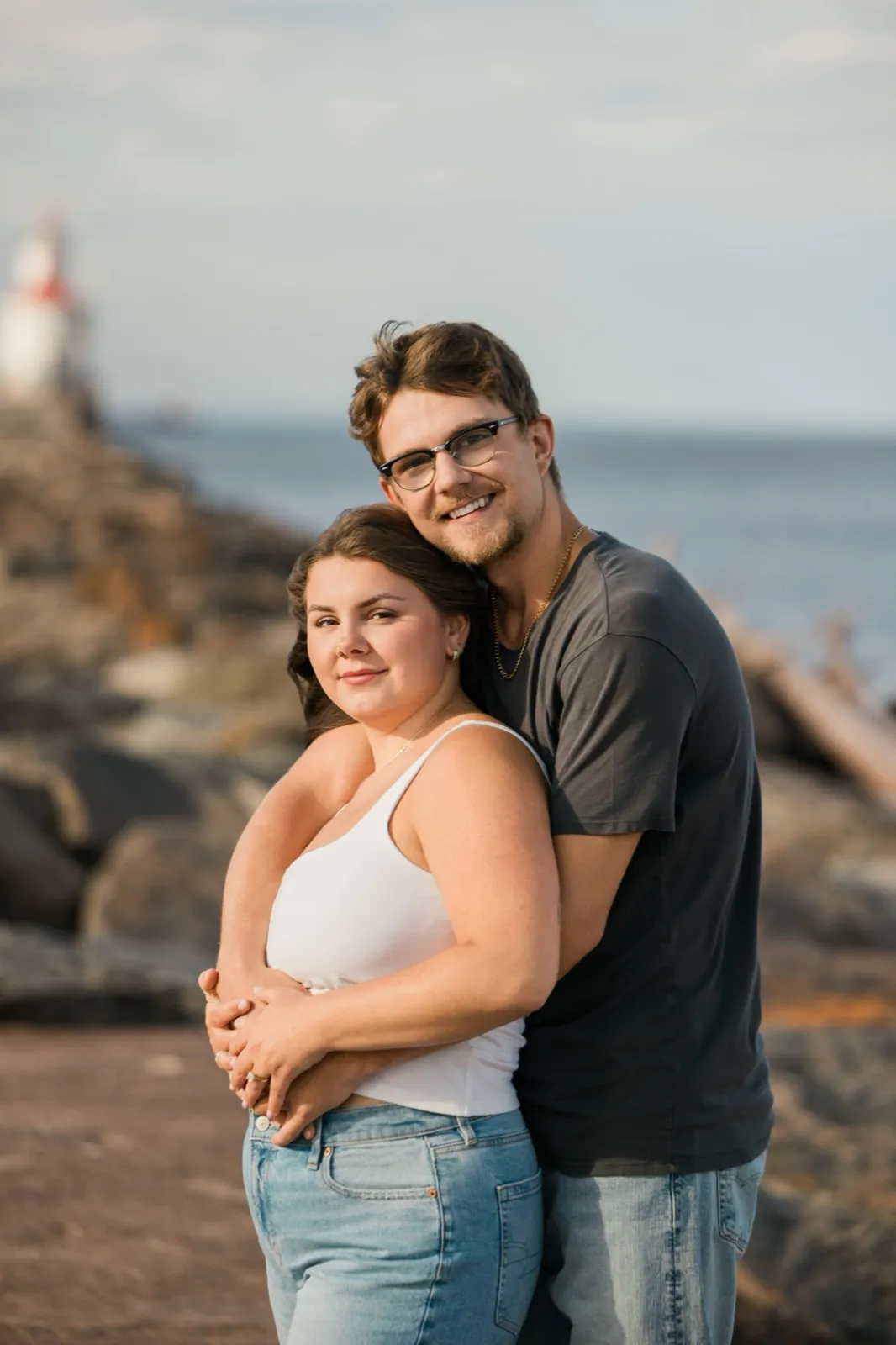Reeden wraps his arms around Jacklyn from behind as they smile for a portrait on the breakwater rocks — Tim Larsen Photography