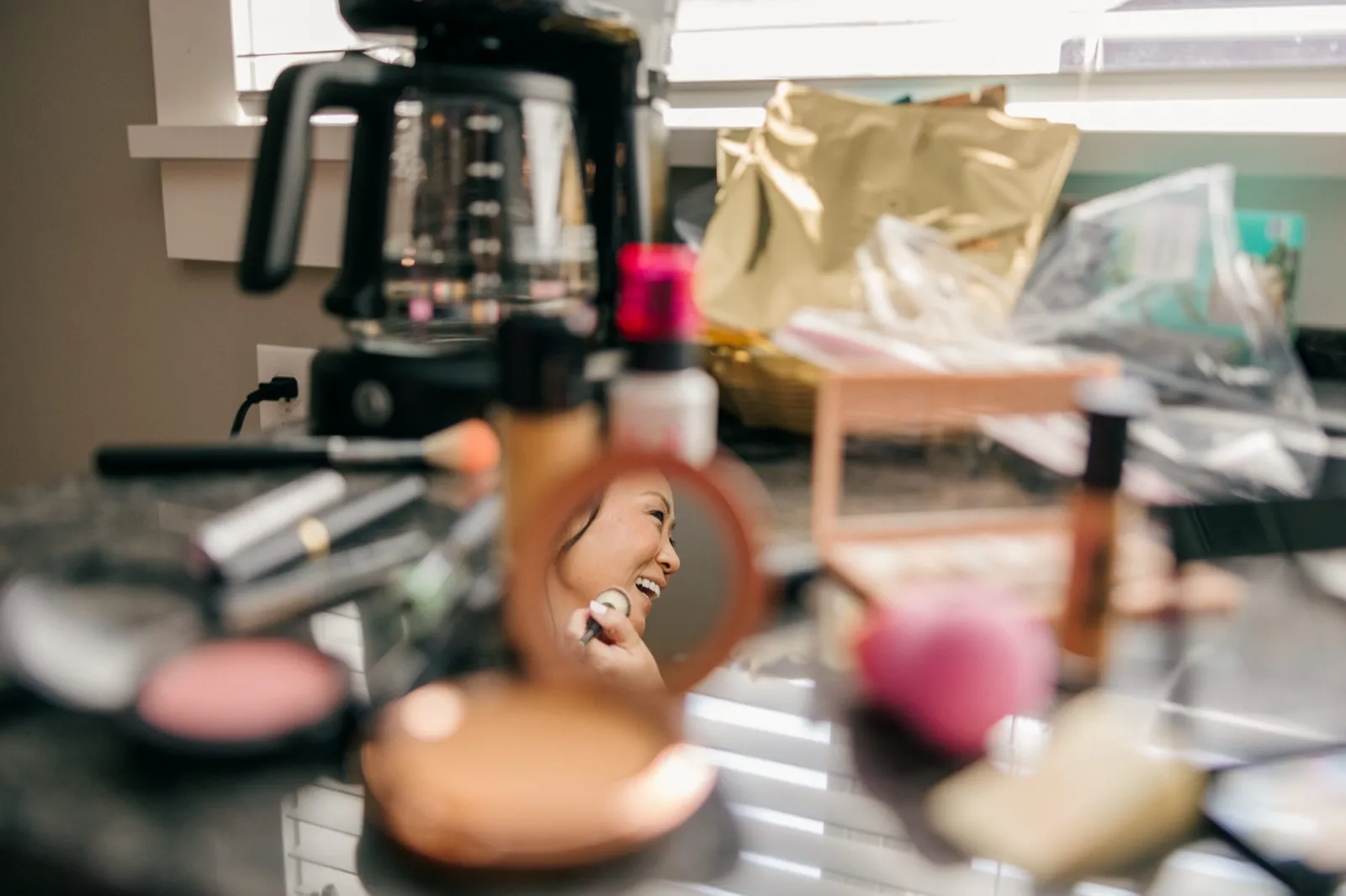 The bride's laughing face appears in a small round compact mirror surrounded by scattered makeup products and a coffee maker on a vanity counter — Tim Larsen Photography, Brainerd Lakes MN