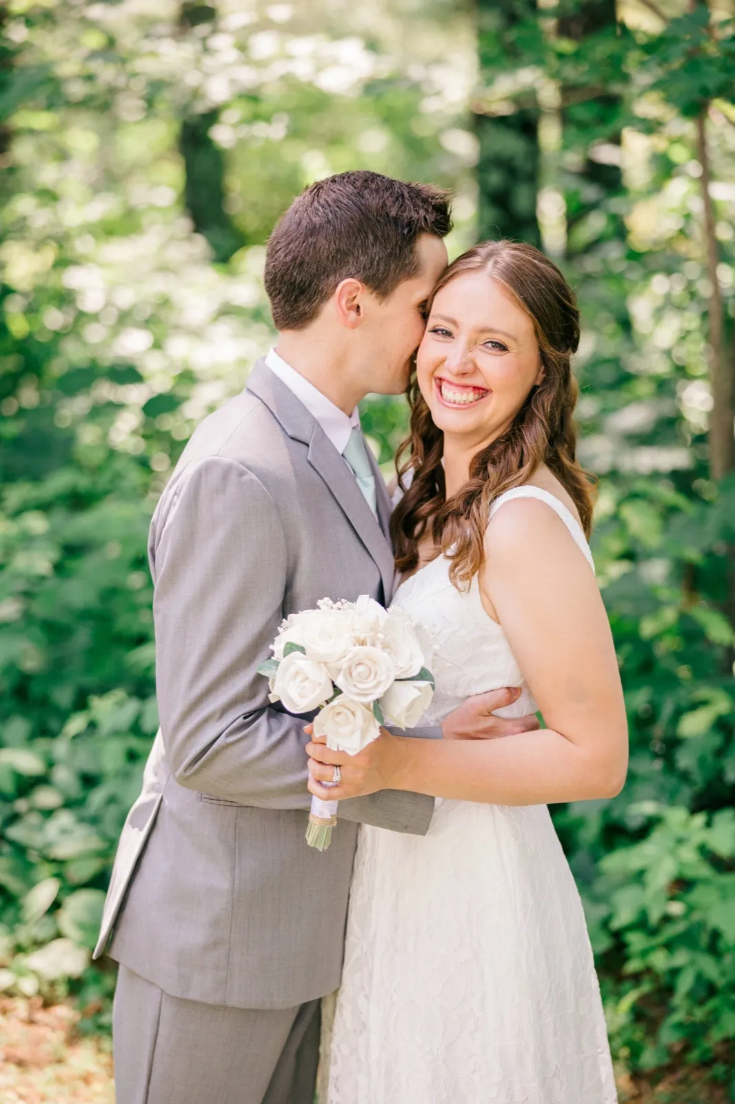The groom leans in to kiss the bride's temple as she laughs broadly toward the camera, holding a white rose bouquet in a sunlit forest clearing. — Tim Larsen Photography, Brainerd Lakes MN