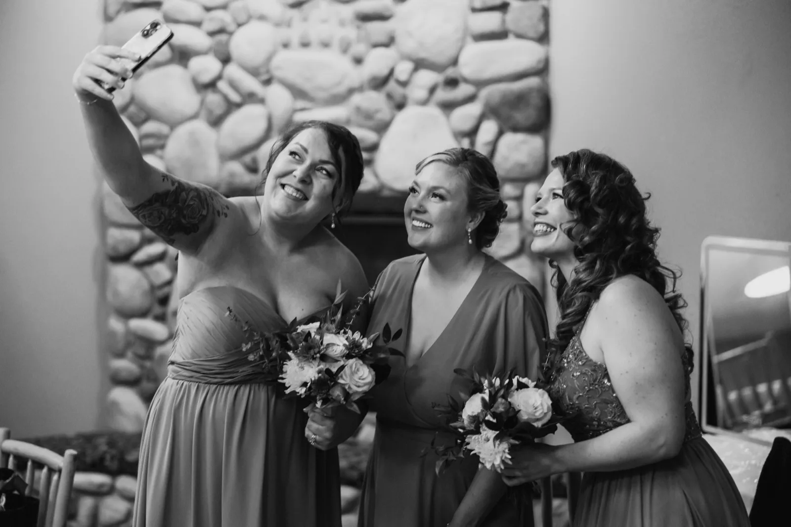 The bride and two bridesmaids holding bouquets lean in for a selfie in front of a stone fireplace at Whitefish Lodge — Tim Larsen Photography, Brainerd Lakes MN