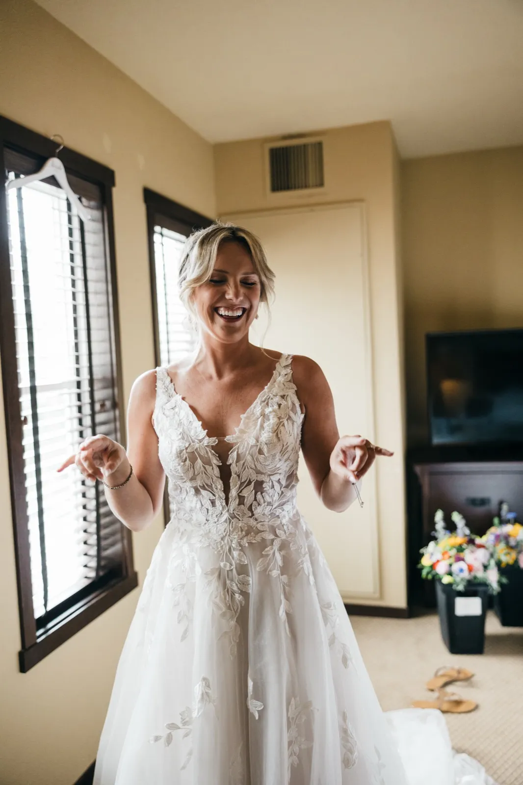 The bride laughs with her eyes closed in her wedding gown, standing in a window-lit hotel suite with bouquets visible behind her. — Tim Larsen Photography, Brainerd Lakes MN