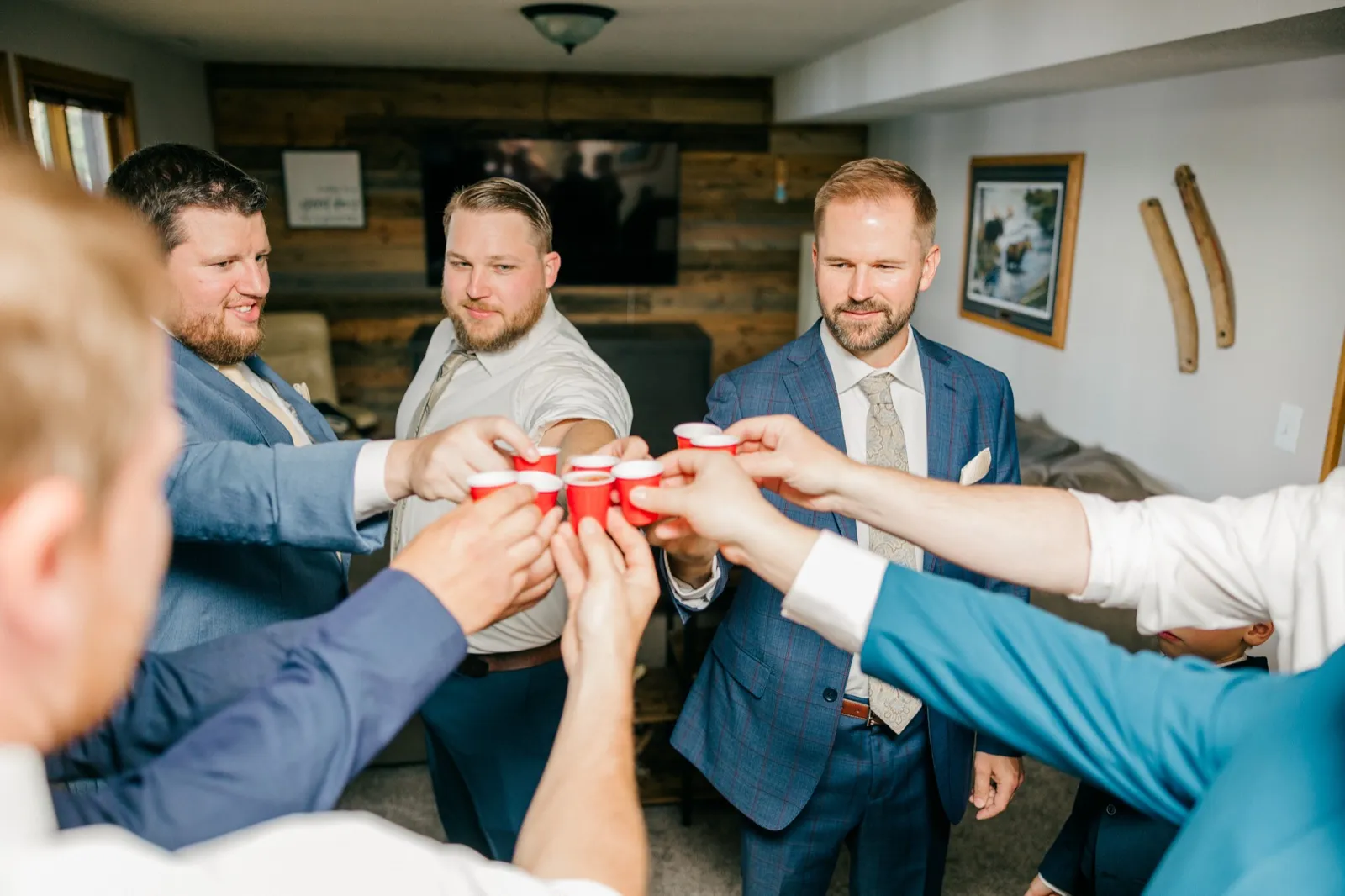 The groom and his groomsmen extend their arms inward to clink small red plastic shot cups together in a wood-paneled cabin lounge — Tim Larsen Photography, Brainerd Lakes MN