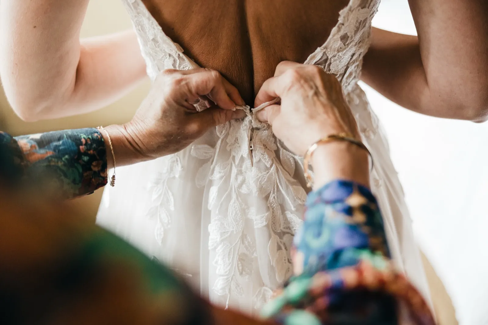 Two pairs of hands, one wearing a floral sleeve and gold bracelet, fasten the lace back of the bride's wedding dress. — Tim Larsen Photography, Brainerd Lakes MN
